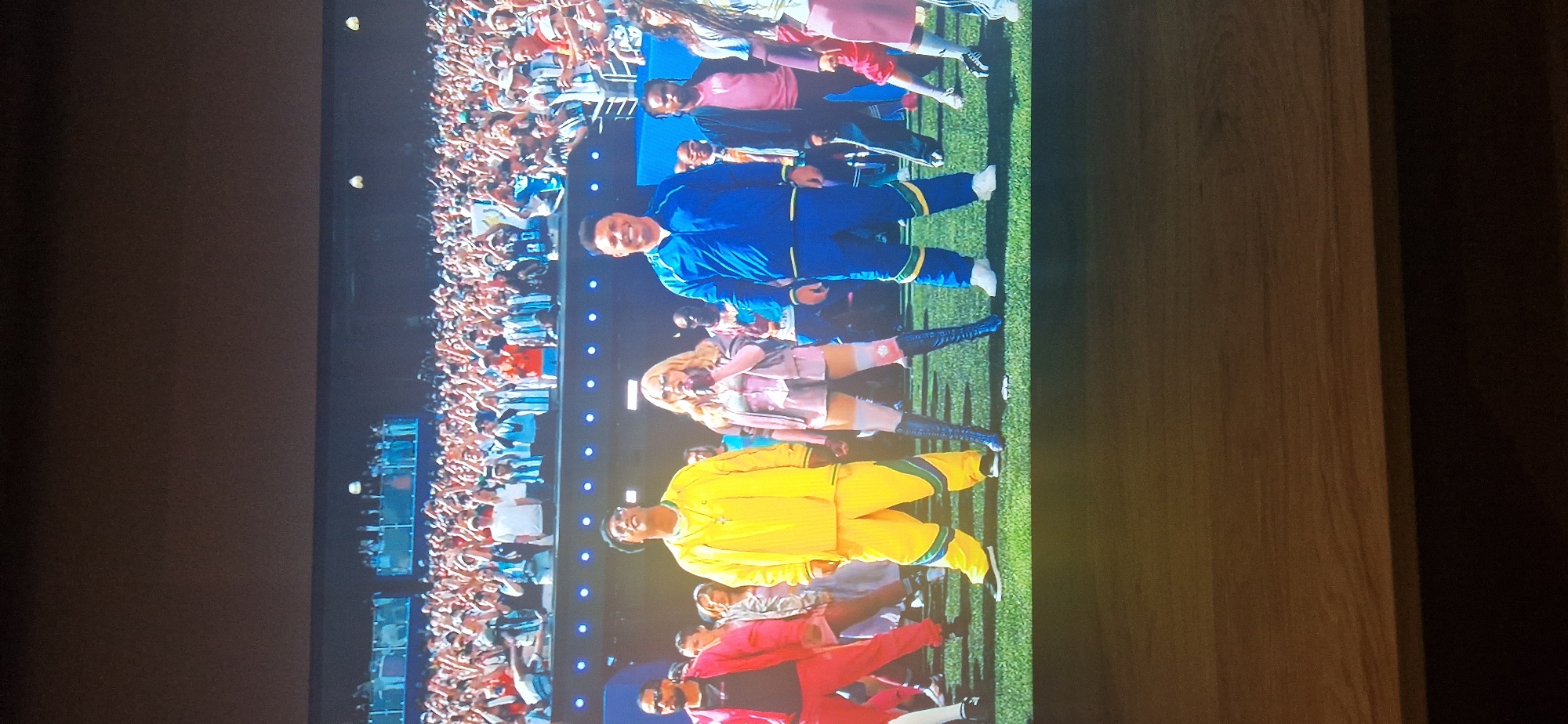 A crowd of people are in the background, most of whom are wearing white and blue striped shirts, suggestive of a soccer match, and many have their hands raised. In the foreground, there are five people standing on a grass-covered surface. From left to right, the first person is a male wearing a bright pink tracksuit and black sneakers. The second person is a male wearing a bright yellow tracksuit with green and blue accents, a necklace, and glasses. The third person is a female wearing a light pink corset top, a pink skirt, and tall, dark blue, lace-up boots. She is holding a microphone. The fourth person is a male wearing a royal blue tracksuit with green accents and white sneakers. The fifth person is a male wearing a dark pink shirt and dark pants. The overall environment appears to be a stadium or outdoor event. There is no visible indication that this location is in Moscow, Russia.