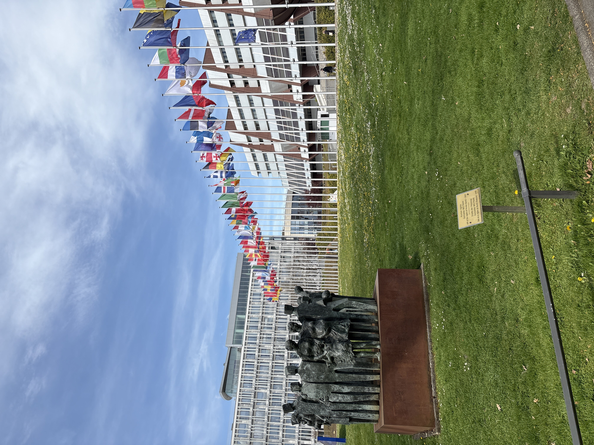 The image shows a photograph taken outside the Council of Europe building in Strasbourg, France. The flags of various nations are displayed in a line, and a bronze sculpture of a group of people stands on a green lawn. A sign near the statue identifies the artist as Gonzalez BELTRAN and states that the work is called “Human Rights”. The sign mentions that the sculpture was a gift from the region of Murcia, Spain, in 2005. The architectural style of the Council of Europe building is visible in the background, and the sky has light cirrus clouds.