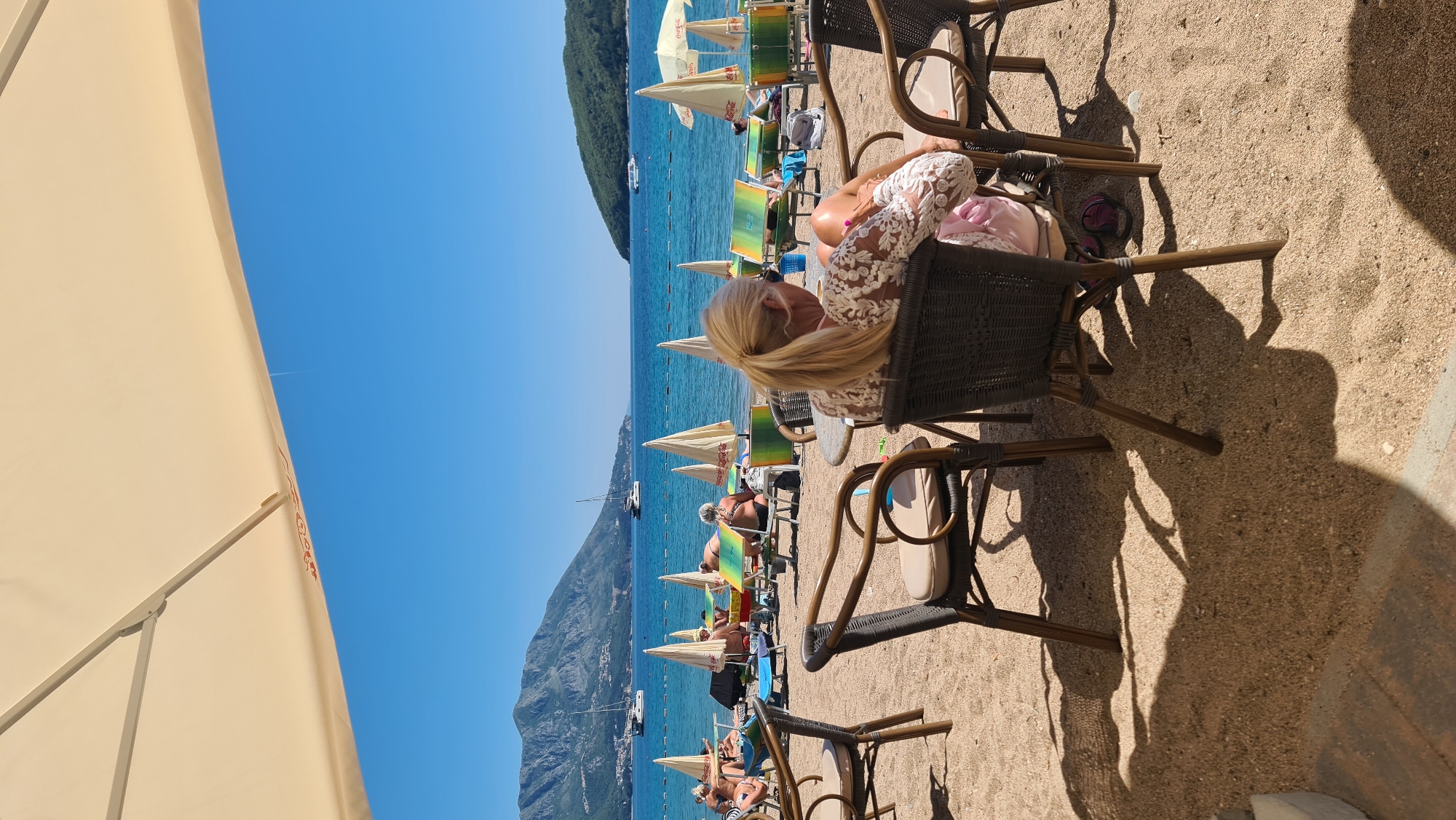 The image depicts a beach scene, likely in Budva, Montenegro. The perspective is from behind a woman with blonde hair in a ponytail, seated on a dark wicker chair at an outdoor cafe. Her back is to the camera, and she is wearing a white lace top. Several other people are visible in the background, relaxing on beach loungers under umbrellas. A mountain is seen in the distance along the coast. The sky is clear and blue, and the sea is calm. A large beige beach umbrella is partially visible on the left side of the frame. The ground is sandy. Other wicker chairs are visible in the foreground.