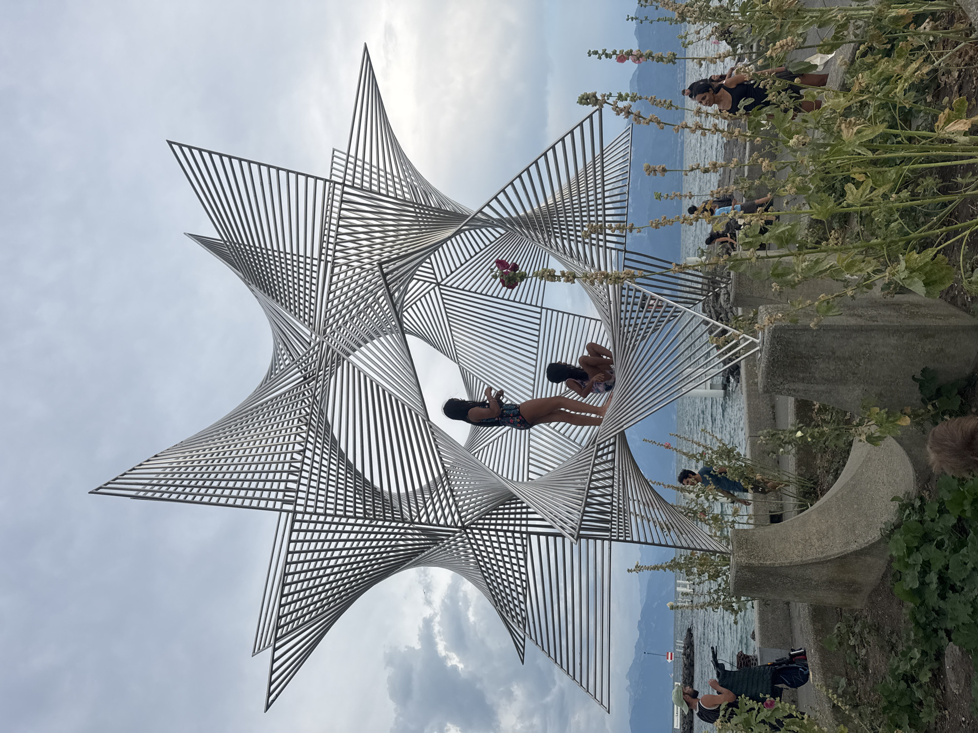 The image depicts an outdoor scene in Lausanne, Switzerland, featuring a large, abstract metal sculpture. The sculpture has a star-like form with intricate geometric patterns created by thin metal bars. Two young girls are interacting with the sculpture; one is standing on it, and the other is sitting. In the background, a body of water, likely Lake Geneva, is visible, with distant mountains and a cloudy sky. Several other people are scattered around the area, some walking along the waterfront and others observing the sculpture. The foreground shows concrete structures and vegetation, including tall plants with flowers and seed pods. A person wearing a green cap is seen to the left, engaged in an activity that appears to be knitting or crocheting. The overall atmosphere is casual and recreational, suggesting a public space like a park or promenade.