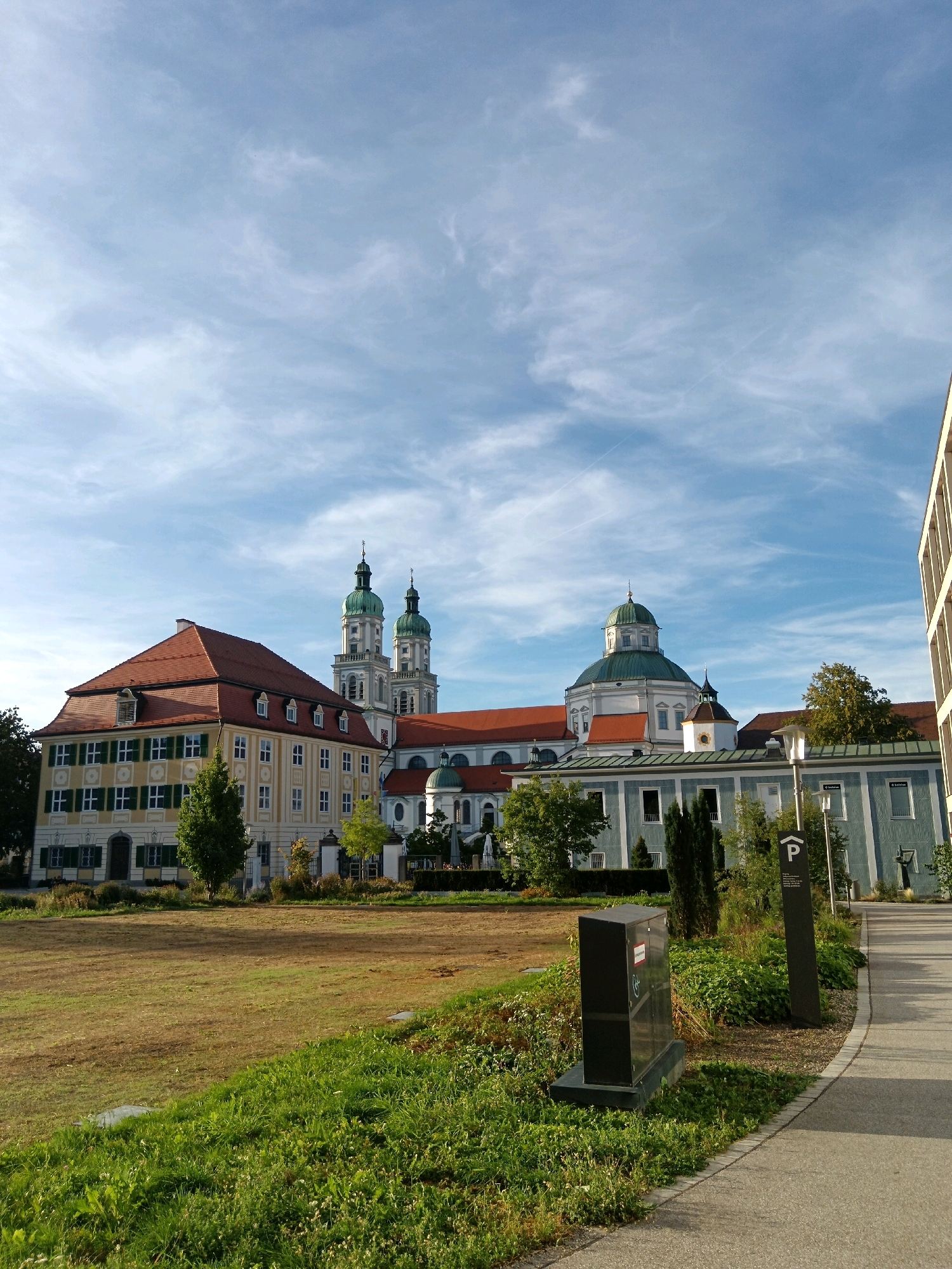 The image shows a view of the St. Lorenz Basilica in Kempten (Allgäu), Germany, on a sunny day with a blue sky and scattered clouds. The basilica, a prominent landmark, is characterized by its two tall, green-domed bell towers and a large central green dome. Adjacent to the basilica are several other buildings with red tiled roofs and light-colored facades.
In the foreground, a paved pathway curves to the right, bordered by a grass verge and lush green foliage. To the left of the pathway, a black electrical box sits on a concrete base within the greenery. A tall, dark grey sign with a white "P" symbol indicating parking is also visible along the pathway.
A large expanse of dry, mown grass occupies the mid-ground, extending towards the buildings. Scattered trees and bushes are present in front of the basilica complex. The overall scene depicts an architectural ensemble set within a well-maintained park-like environment under a bright sky.