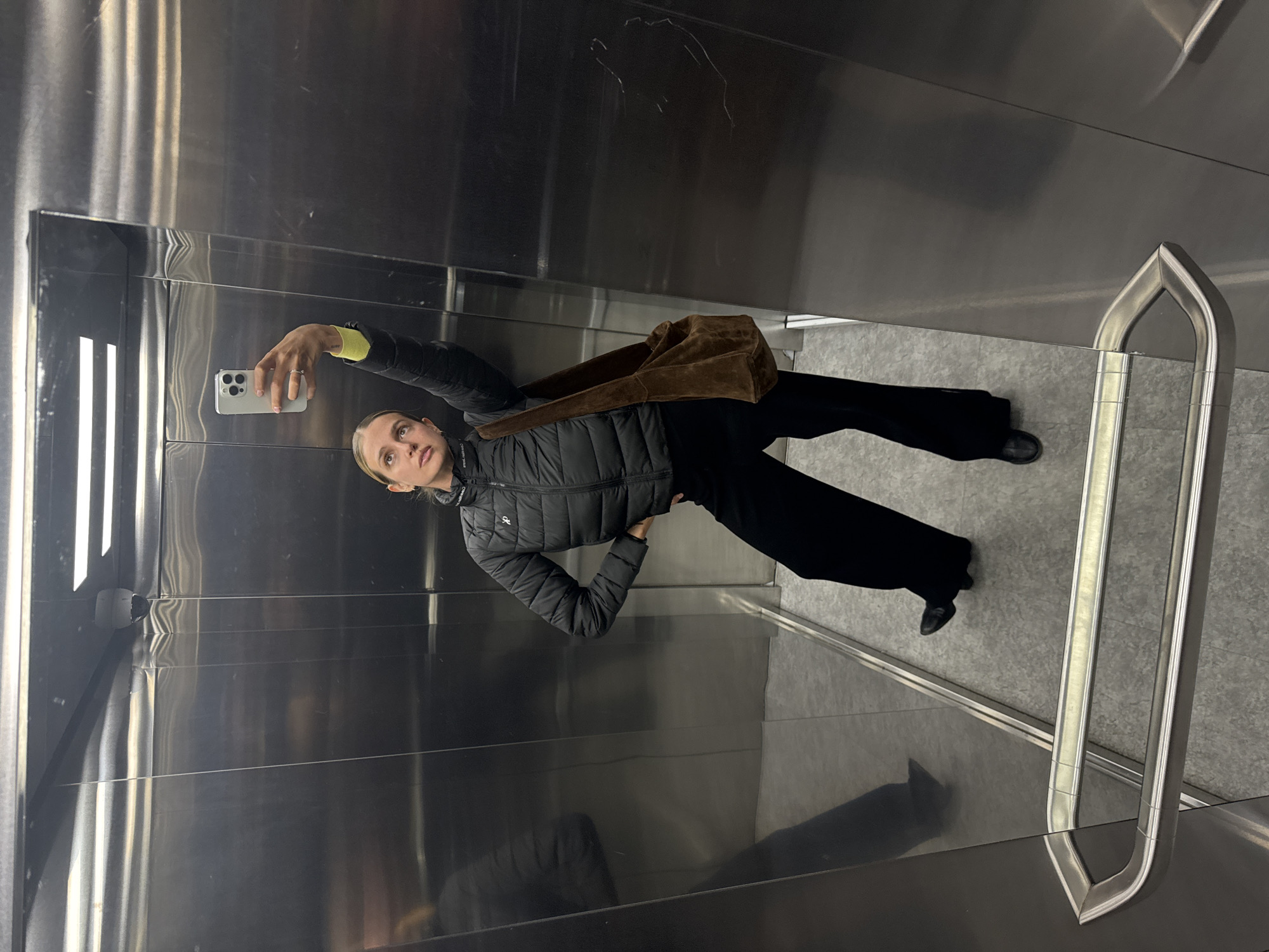 A young woman takes a selfie in a mirrored elevator. She is wearing a black puffer jacket with a yellow sleeve cuff visible, black pants, and black shoes. A brown bag rests on her shoulder. The elevator has shiny metal walls and a gray tiled floor. A silver handrail is visible. The location is Unknown, Unknown.