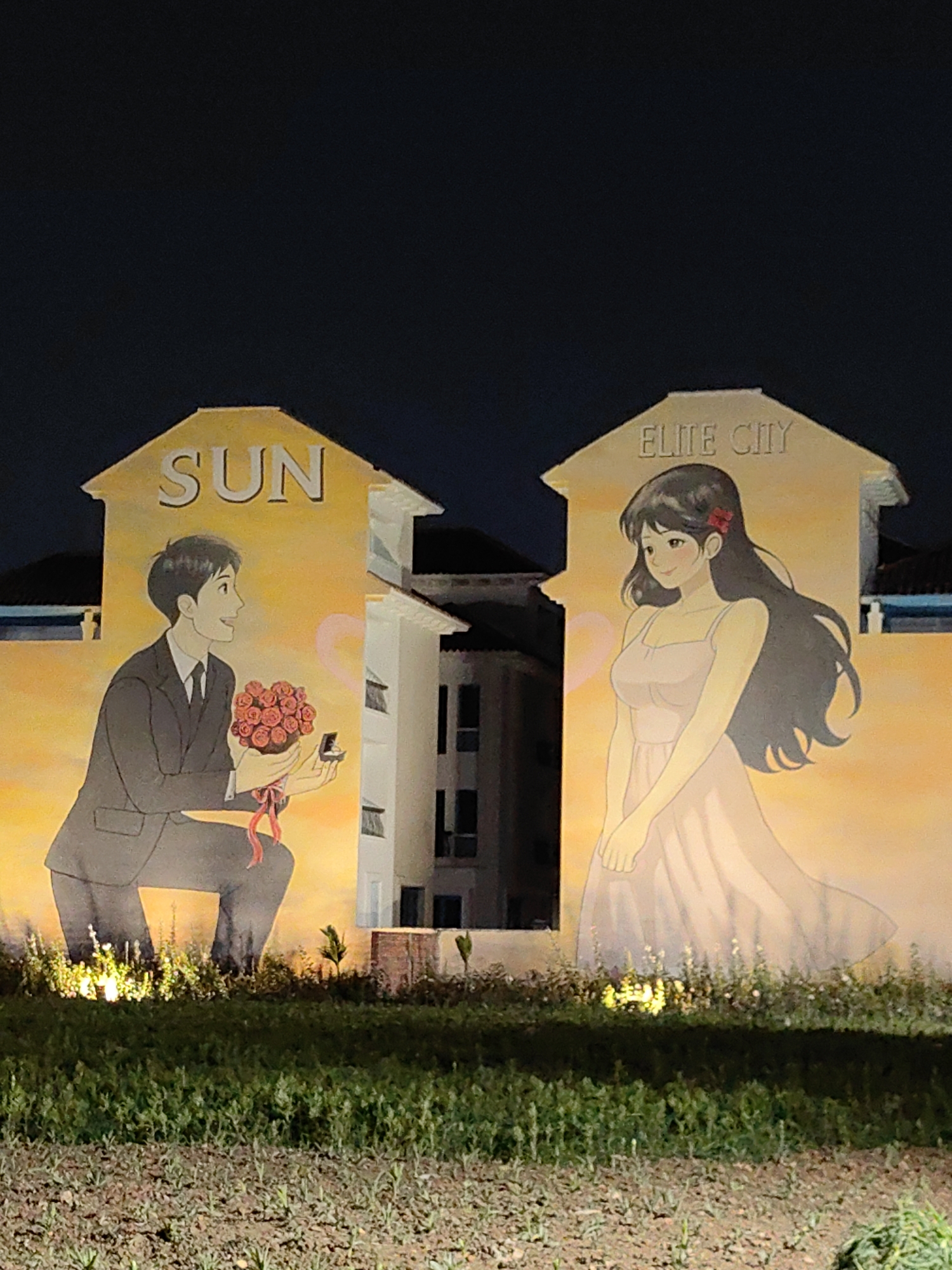 The image captures a scene at night in Bai Chay Ward, Vietnam. Two separate buildings are visible, each adorned with murals depicting a romantic narrative. The building on the left has the word "SUN" emblazoned on its roof. Below, a mural depicts a man in a suit kneeling, holding a bouquet of red roses and a ring box, seemingly proposing. The adjacent building, separated by a visible portion of another structure, features the text "ELITE CITY" at the top. A mural below shows a woman in a light-colored dress, looking at the man. The images are rendered in an anime style. Both buildings appear to be painted a yellowish-orange color in the mural areas. The ground in front of the buildings is covered with grass and small plants. The scene is illuminated by some artificial lights, with a dark night sky as the backdrop.
