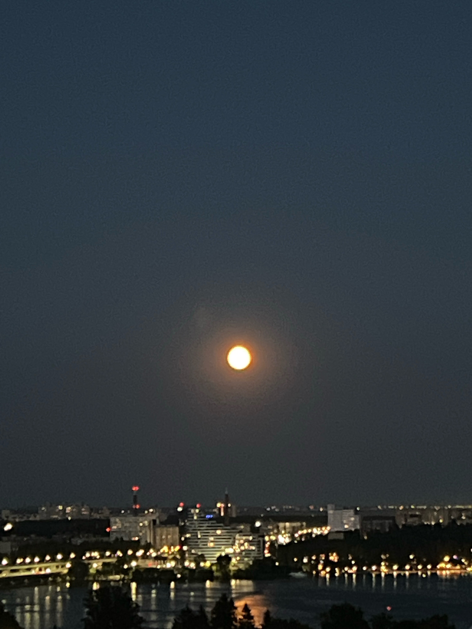A full moon hangs in the dark night sky over a city skyline. The moon is a bright, glowing orb with a reddish-orange hue. Below, the city is illuminated by countless lights, reflecting off a body of water in the foreground. Buildings of various heights and designs are visible, with some appearing modern and others older. A bridge with lights runs across the water. The scene suggests a peaceful evening view of an urban area at night. The location is Novaya Usman, Russia.