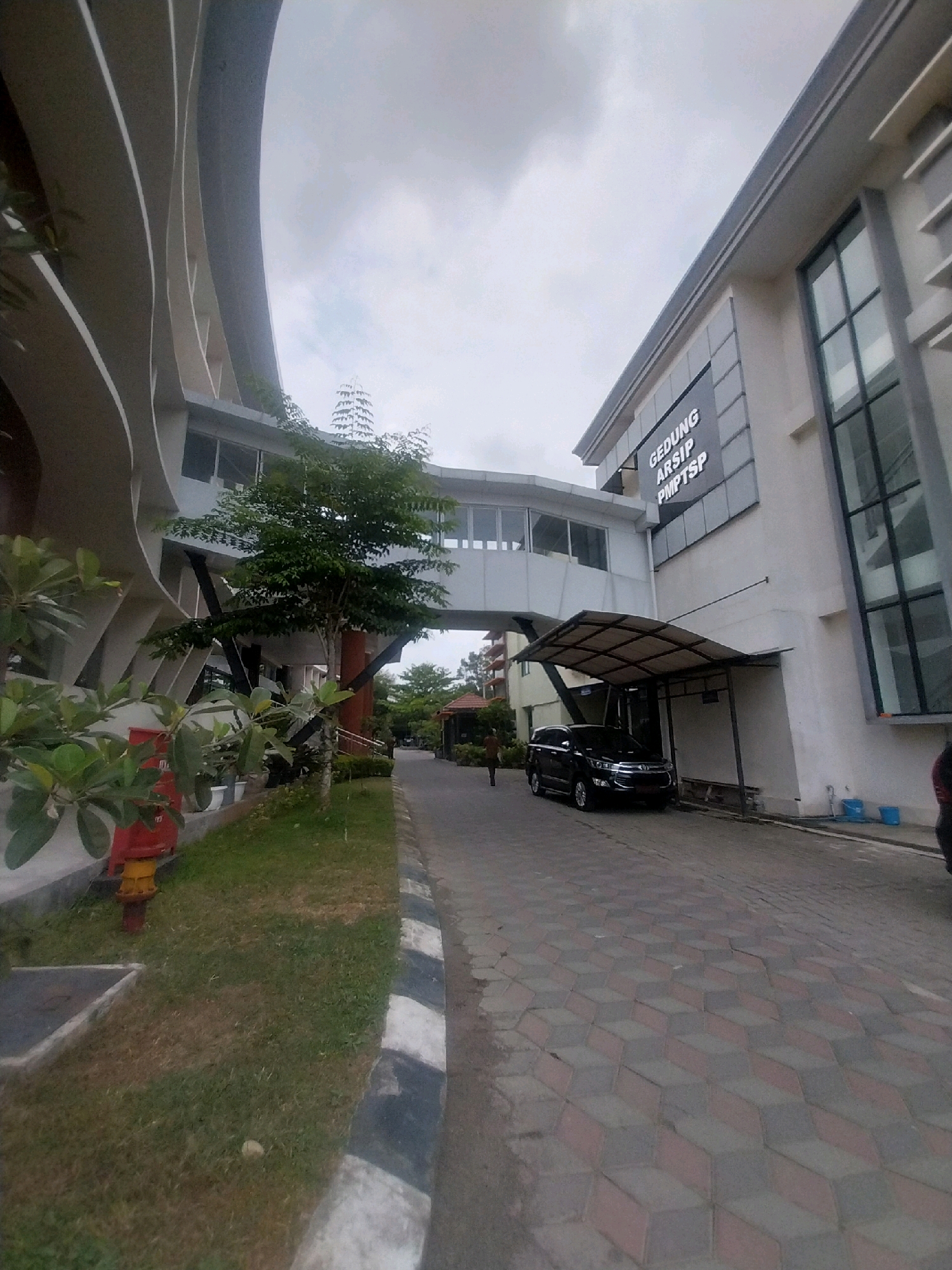 The image depicts the exterior of the GEDUNG ARSIP PMPTSP building in Sleman, Indonesia. The building features a modern architectural style with a curving facade on the left, juxtaposed with a more linear structure on the right. A skybridge connects the two sections of the building at an upper level. In front of the right section, a black SUV is parked under a canopy. The ground is paved with interlocking hexagonal and rectangular blocks that create an illusion of depth. To the left of the paved area, a grassy lawn is visible, with a red fire hydrant and some potted plants. The sky is overcast. A person is partially visible walking on the paved area in the distance.