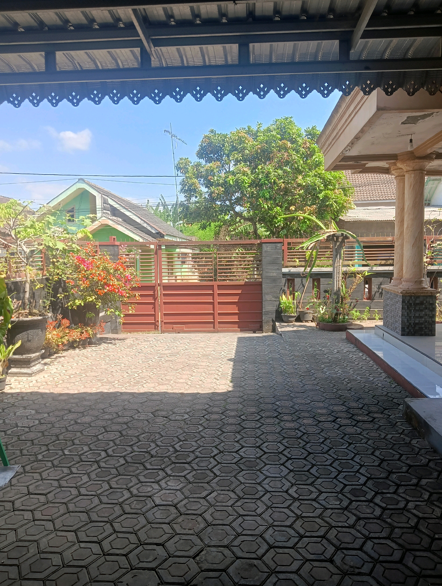 The image captures a view of a paved yard and the front of a residential property. The yard is covered with interlocking hexagonal pavers. In the background, a brown metal gate is visible, separating the yard from a street or driveway. Beyond the gate, there is a house with a light green exterior and a dark grey roof. A large, leafy green tree stands to the right of the gate, casting a shadow on the yard. On the right side of the image, part of a building's facade is visible, featuring decorative columns and a light-colored wall. Several potted plants, including one with bright orange flowers, are placed along the left side of the yard. The sky is clear and blue with a few white clouds. The scene appears to be a typical residential setting in Kromengan, Indonesia, likely captured during daylight hours. There are no people or animals visible in the image.