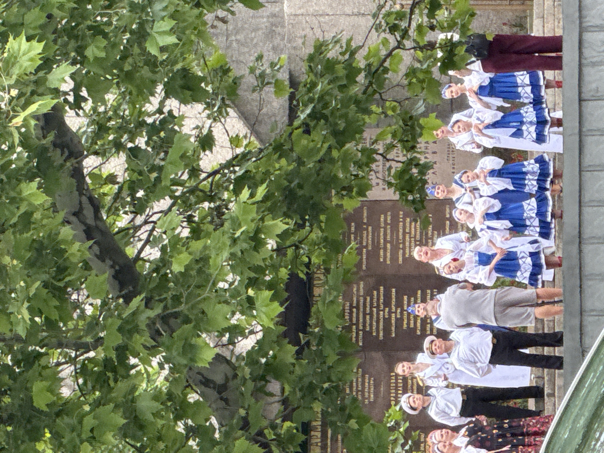 This detailed image captures an outdoor cultural performance or ceremony set against a stone monument partially obscured by dense green foliage. The scene is illuminated by ambient daylight, suggesting an overcast sky or filtered sunlight. In the background, a large, light-colored stone wall or monument is visible, featuring multiple dark rectangular plaques. These plaques bear inscriptions, primarily in Cyrillic script, with words like "ОБОРОНЫ" (Defense) and "ФРОНТОВАЯ" (Frontline) discernibly suggesting a memorial, likely for military or historical figures. The monument's base appears to be a stepped structure. A large tree with broad, green leaves and thick branches dominates the upper portion and mid-left of the image, casting dappled shadows and partially obscuring the monument. On a raised platform or steps in front of the monument, a diverse group of people is assembled. On the far left, a woman in a dark, patterned dress, with floral or abstract motifs in red and white, is partially visible. Next to her stands a woman in a white long-sleeved folk dress with red detailing on the collar and cuffs. In the central-left area, two men are dressed in white sailor-style uniforms: white long-sleeved shirts, black trousers, and white sailor caps. One man on the left looks upwards and to the left, while the other, slightly to the right, stands with his hands clasped behind his back, facing forward. Behind them, a tall woman in a long white coat or uniform with gold accents, her hair pulled back, looks towards the right. Further to the right, a man in a light grey t-shirt and light grey shorts stands with his back mostly to the viewer, looking towards the right side of the group. He has a dark object, possibly a camera or small bag, attached to his left hip. On the right side of the platform, a group of approximately seven women and girls are uniformly dressed in blue and white folk costumes. These outfits feature white puffed sleeves, blue bodices, and full blue skirts with prominent white geometric patterns. They also wear white headscarves or bands. Many are standing with their hands clasped together at chest level, indicative of a performing or singing stance. One woman in a long white coat or dress stands behind this group, looking forward. Another woman with a blue cap and white uniform is partially visible behind the group. On the far right, a woman wearing dark maroon trousers