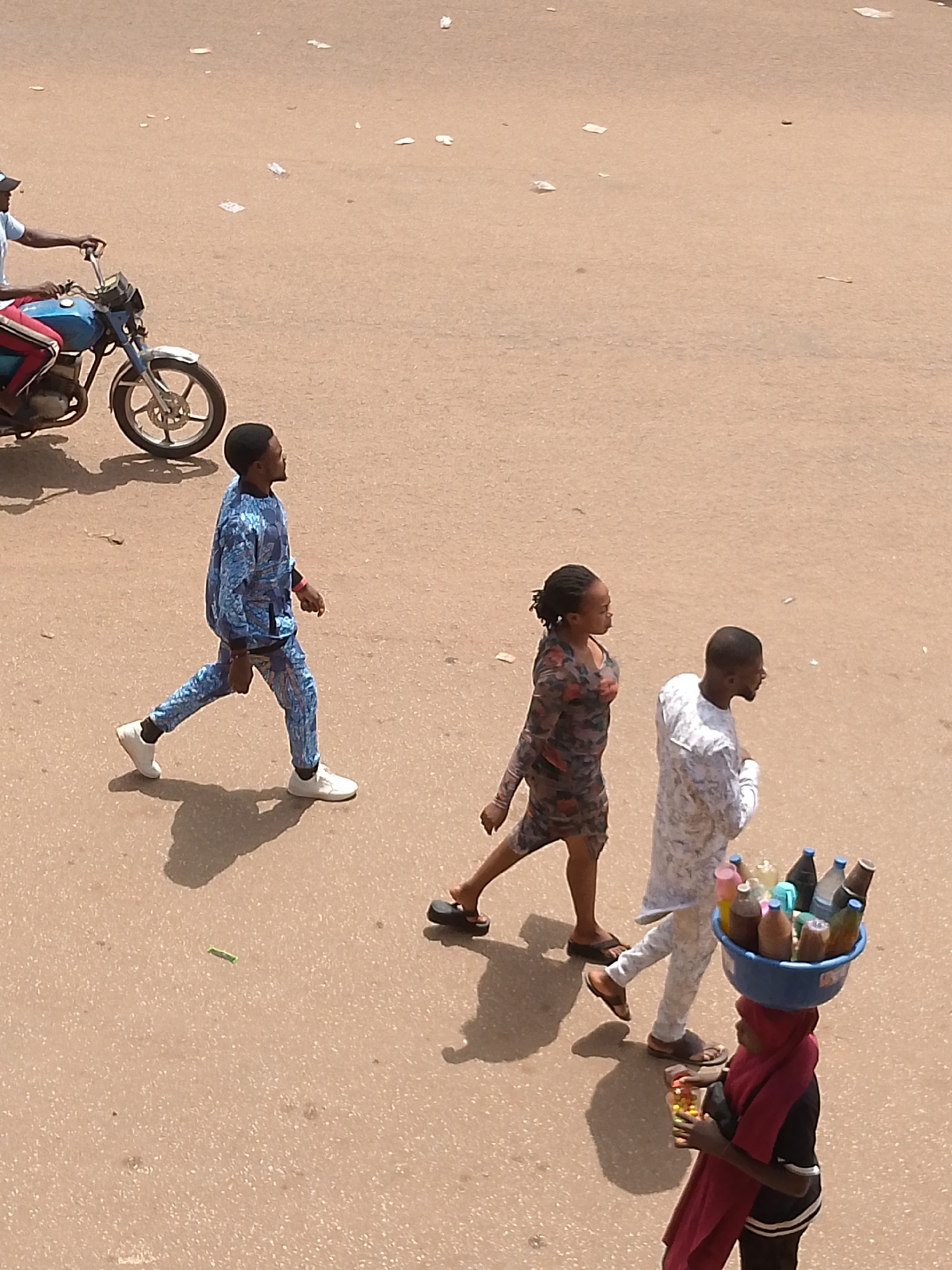 The image, likely taken in Bukuru, Nigeria, shows an overhead shot of a street scene. A man on a blue motorcycle is on the left edge of the frame. Three pedestrians are walking rightward in the middle of the image: a man in a patterned blue suit, a woman in a patterned dress, and a man in a patterned white outfit. In the lower right, a woman carries a blue basin filled with bottles on her head. She wears a red scarf, a black top, and black pants. There is trash scattered across the street.