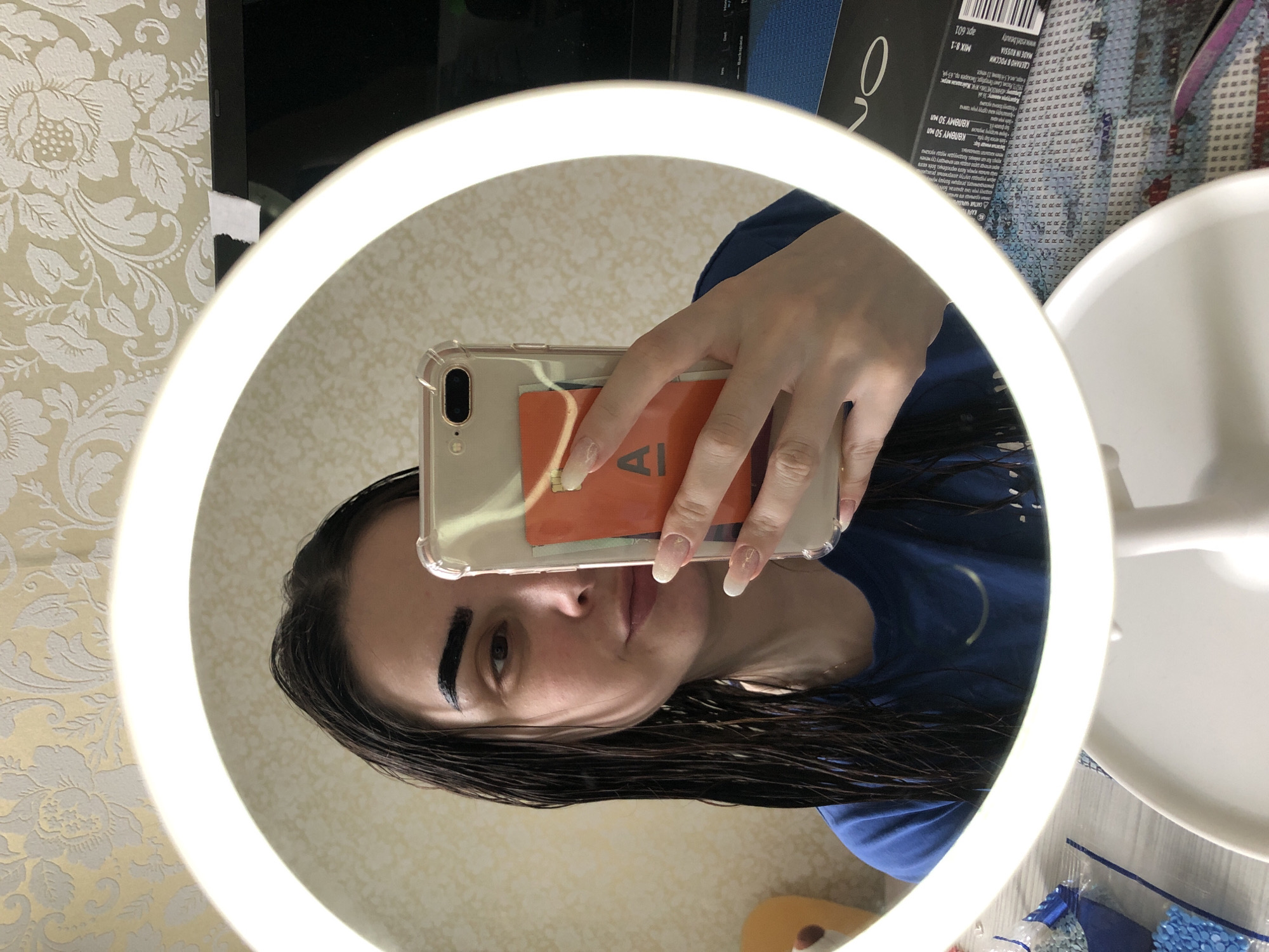 A person is taking a selfie in a lighted mirror. The mirror is surrounded by a ring light. The person is holding a smartphone with a clear case and an orange card visible behind it. Their hair is dark and wet. The background includes patterned wallpaper, a white surface, and a cosmetic box. The OCR text indicates the product is made in Russia and includes the location: 195371, Russia, Saint Petersburg, Piskarev pr. 63-y. There is no information about the city of Noviy.