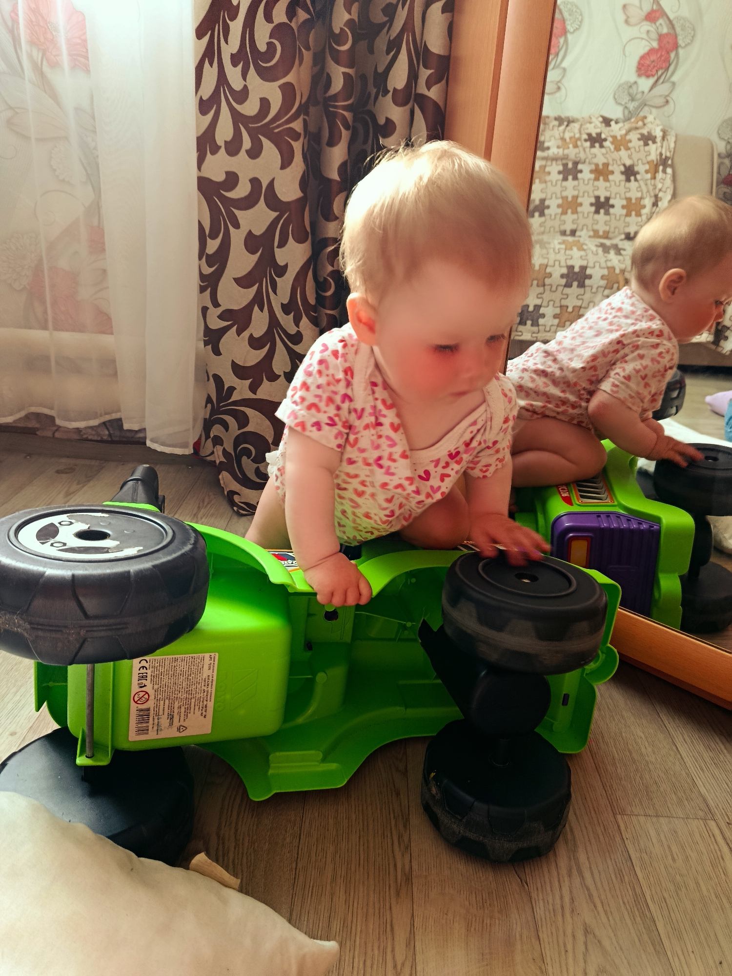 A baby with fair skin and light-colored hair sits on a green toy vehicle with black wheels. The baby is wearing a white onesie with small pink heart patterns. In the background, partially obscured by a wooden frame, is a reflection of the baby and part of the room including a couch. The location is Фоки, Russia. The surface beneath the baby is a light-colored wooden floor. A brown and beige patterned curtain hangs in the background to the left. A white curtain with a floral pattern is visible near the brown curtain. A white object is in the lower-left.