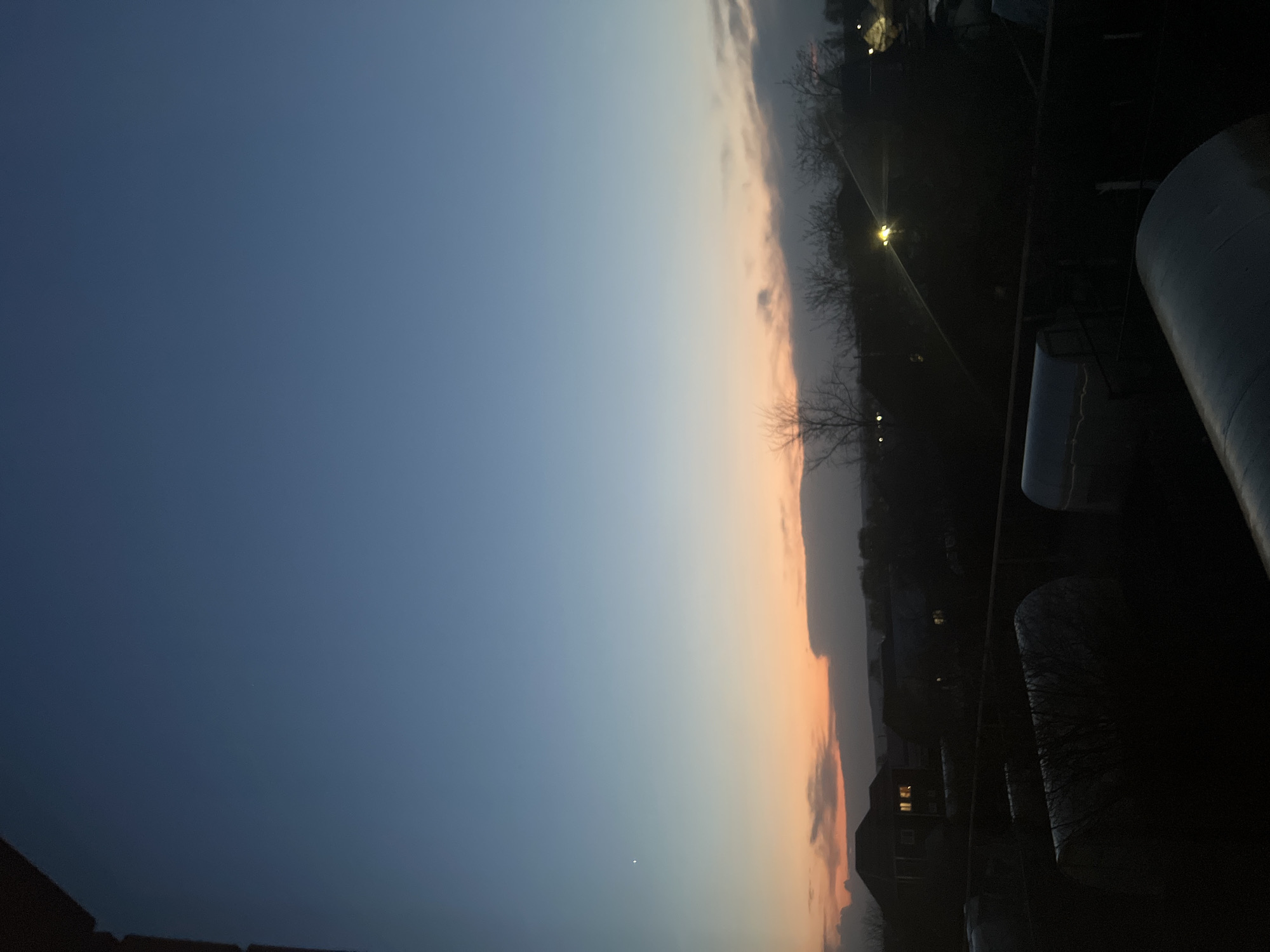 The image shows a dusk or dawn scene. The sky transitions from a dark blue at the top left to an orange glow near the horizon. There are some clouds along the horizon. In the foreground, the silhouette of a structure, possibly a building or balcony, can be seen. In the distance, lights suggest a town or city environment with some buildings and streetlights.
Without additional visual details or context, it's impossible to definitively confirm if the location is Votkinsk, Russia.