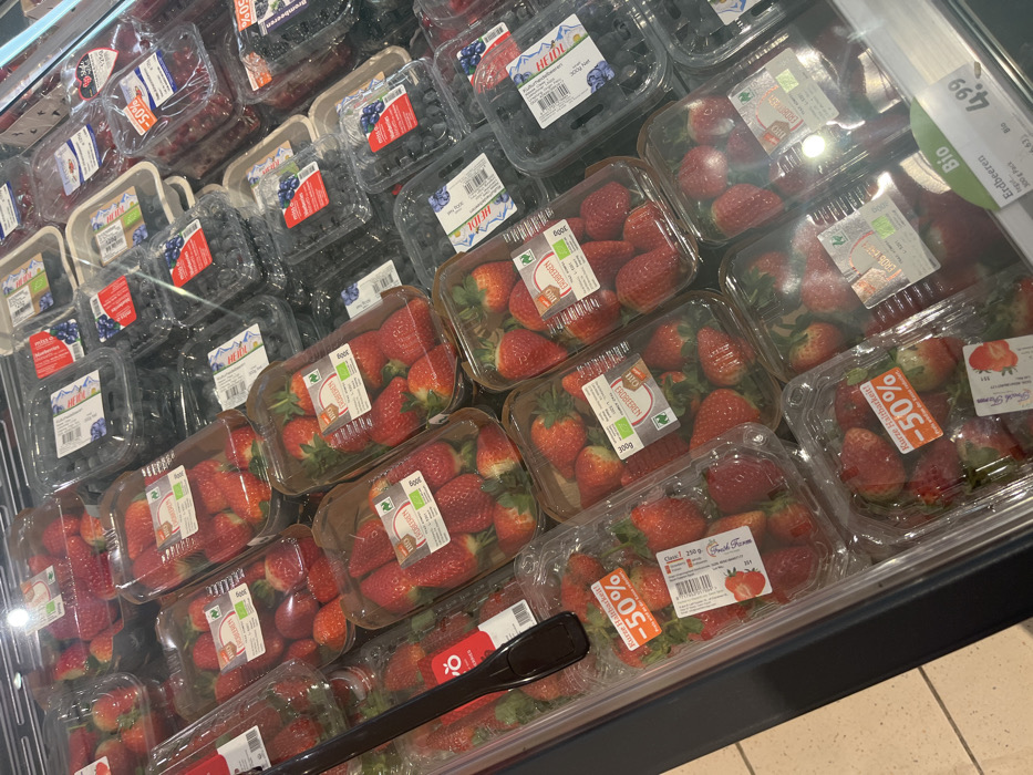 This media file captures a close-up view of a refrigerated produce display in a supermarket located in Bamberg, Germany. The primary subjects are numerous plastic containers filled with fresh berries, mainly strawberries and blueberries.
The strawberries are packaged in clear plastic punnets, many of which carry labels indicating "ERDBEEREN" (strawberries) and a weight of "300g." Several of these labels also feature a "BIO" (organic) certification logo. A prominent detail is the presence of red and orange stickers on some strawberry containers, advertising "Kurze Haltbarkeit!" (short shelf life!) and a "50%" discount. Brand names such as "HEIDL" and "Fresh Farm" are visible on the packaging.
Blueberries are also displayed in clear plastic containers, with some appearing to have a molded fiber base inside, also bearing "HEIDL" and "BIO" labels. In the upper right section of the image, a larger white price tag is visible, indicating "Bio ERDBEEREN" in a "300g Pack" for "4.99," with a smaller number "6.63" likely representing a comparison price. The items are neatly arranged and stacked in the brightly lit display case, with reflections visible on the plastic surfaces. The scene depicts a typical retail environment for fresh produce, implying an activity of grocery shopping. No people are visible in the image.