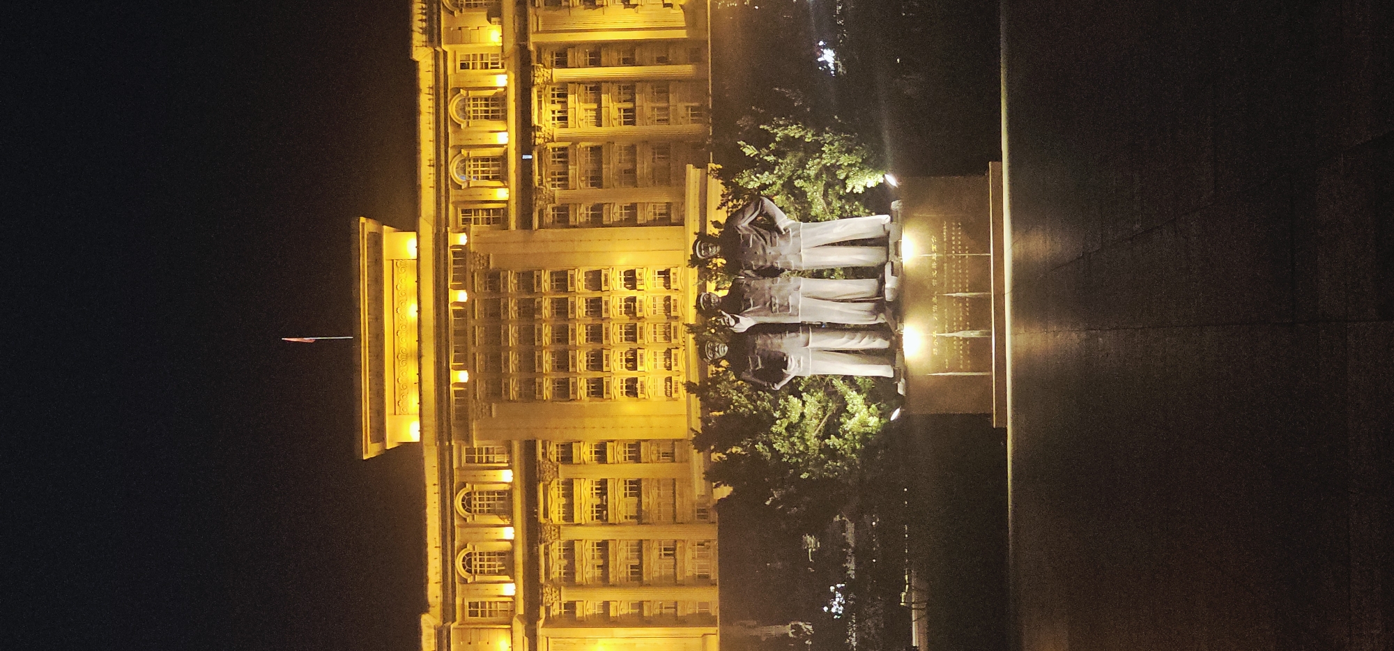 The photograph captures a nighttime scene in Beibei, China, featuring a monument in the foreground and a grand, brightly lit building in the background. The monument consists of three full-body bronze statues of men standing side-by-side on a stone pedestal. The statues appear to be of historical figures, all wearing similar attire: jackets, shirts, and trousers, with one figure in the center pointing upwards. The pedestal has inscriptions, but they are not clearly legible in the image. Behind the monument, lush green trees are illuminated by spotlights. Dominating the background is a large, multi-story building, bathed in a warm, golden light. The building exhibits classical architectural elements, including columns and rows of windows. A small flag is visible atop the building. The foreground consists of a paved area, reflecting the light from the monument and the building. The overall impression is one of solemnity and historical significance.