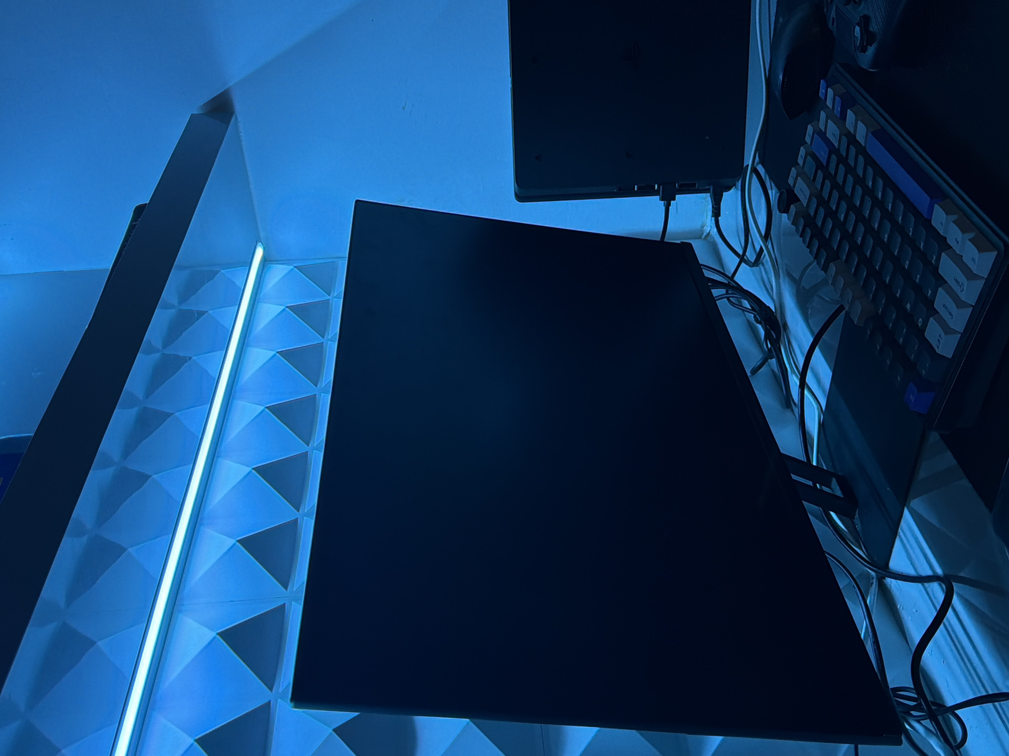 The image shows a gaming setup in a room, illuminated by blue LED lighting. There are two monitors, one large one in the foreground and a smaller one to the right. A keyboard and mouse are on a desk in front of the monitors. The walls have a geometric texture and are lit by a strip of blue light. There are no people or animals visible in the image. The scene is a typical home or office setup for a gamer or computer user. The location in Wonoayu, Indonesia is not discernible from the image.