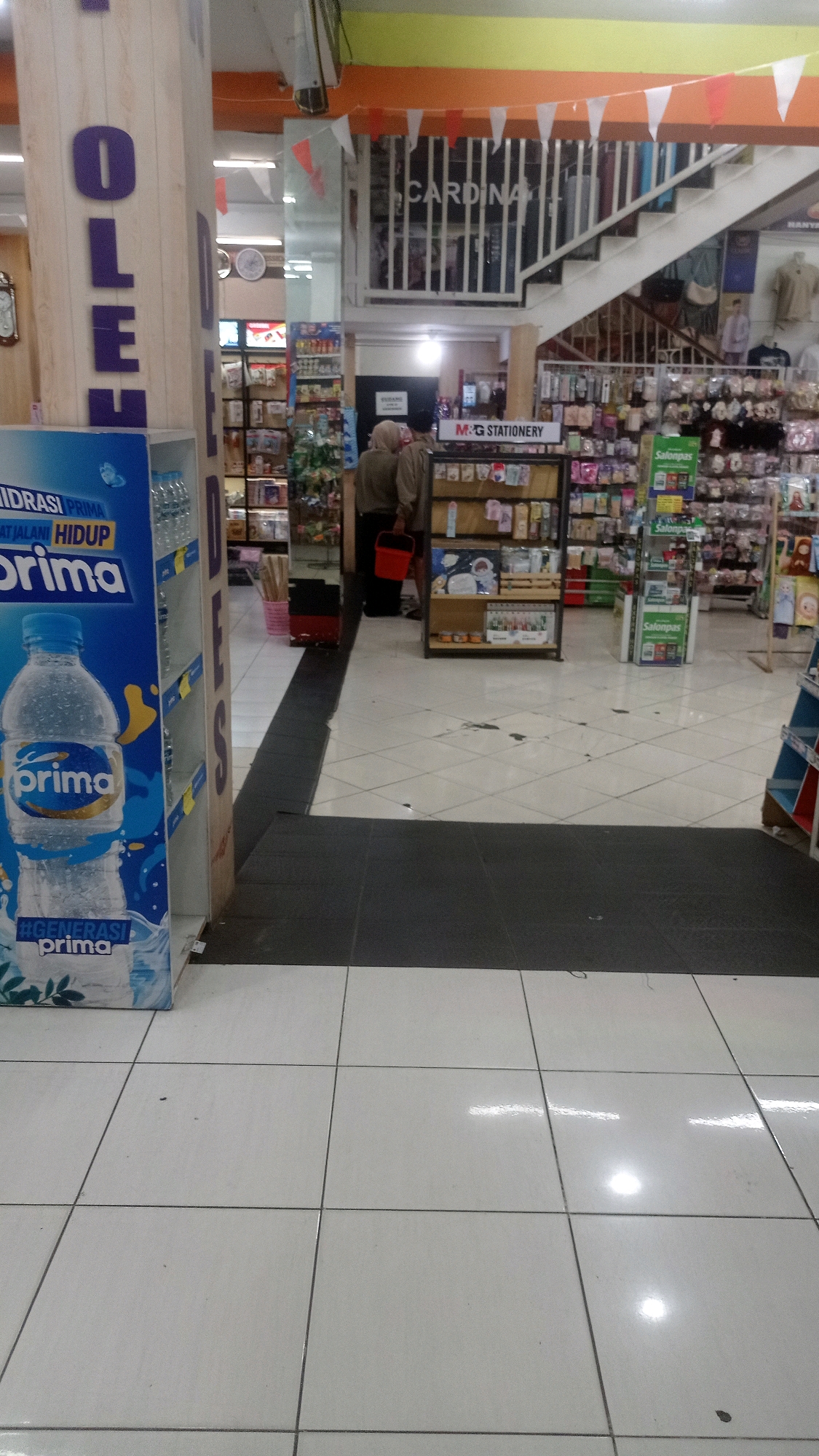 The image depicts the interior of a retail establishment, likely a supermarket or a department store, located in Mulyoagung, Indonesia. In the foreground, a large advertisement for "Prima" bottled water is visible on the left side of the frame, featuring a bottle of water and branding. To the right of the advertisement, a pillar with the letters "OLEH" vertically printed in purple is present.
Further into the store, aisles of merchandise are organized on shelves. In the middle ground, two individuals, one appearing to be a woman in a hijab and another person in casual clothing, are seen walking. The woman is carrying a red bucket. A prominent sign that reads "M&G STATIONERY" is displayed above a shelf unit stocked with various stationery items.
In the background, a staircase leads to an upper level. A sign with the word "CARDINA" is visible on the railing of the staircase. The store is brightly lit, with fluorescent lighting illuminating the space. The floor is tiled with a checkered pattern of white and grey tiles. Various products are displayed throughout the store, suggesting it is a well-stocked retail environment. The overall impression is that of a typical Indonesian commercial interior.
