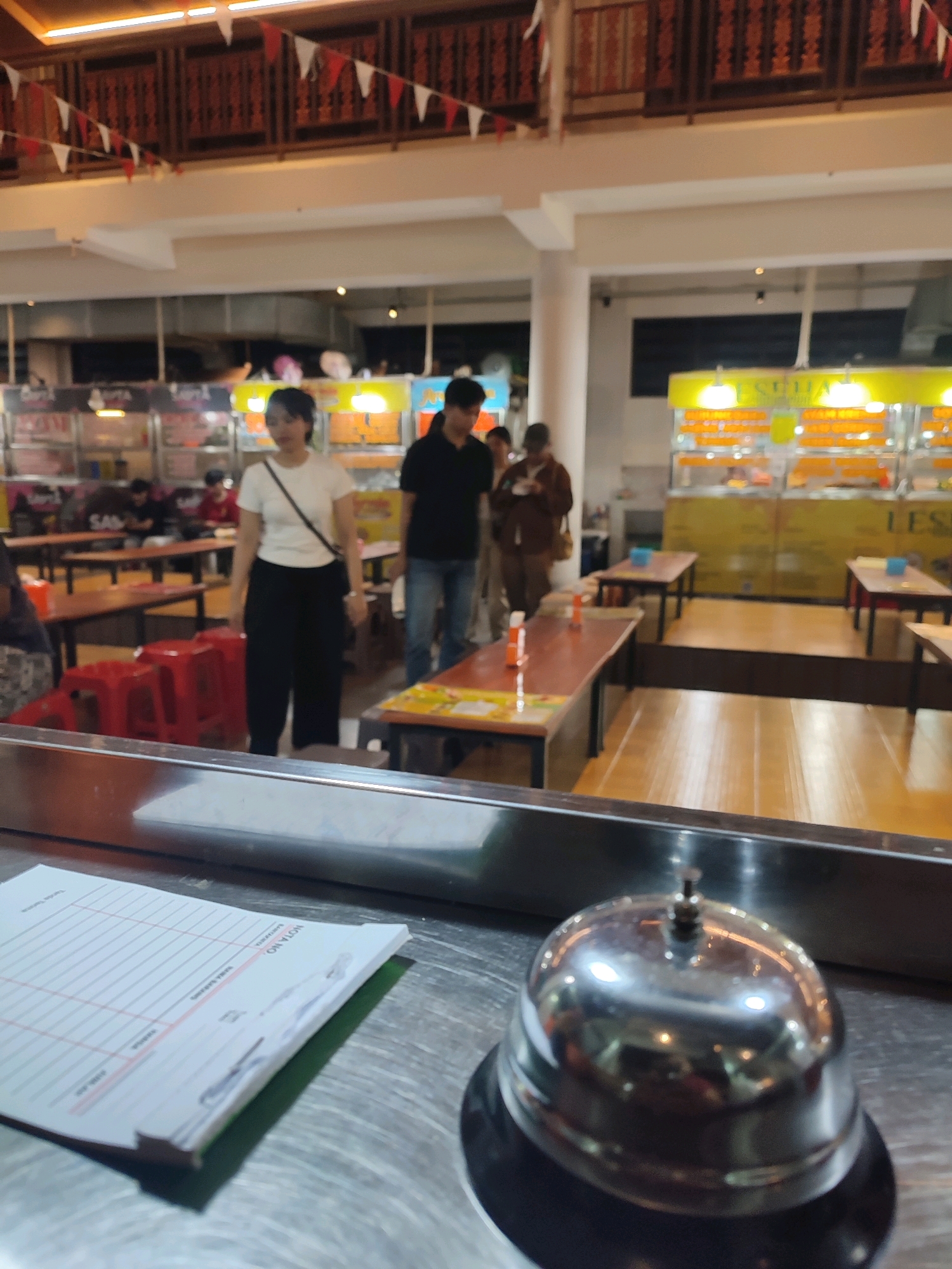 The image depicts a food court in Yogyakarta, Indonesia, characterized by its brightly lit food stalls and communal dining tables. In the foreground, a silver service bell sits on a metallic counter, accompanied by a stack of order forms, suggesting a busy service environment. The background reveals several food vendors with illuminated signs and displays showcasing their offerings. People are visible in the middle ground, some standing and interacting near the food stalls, while others are seated at tables, indicating a casual dining atmosphere. The overall scene suggests a typical bustling evening at a popular food court in the city.