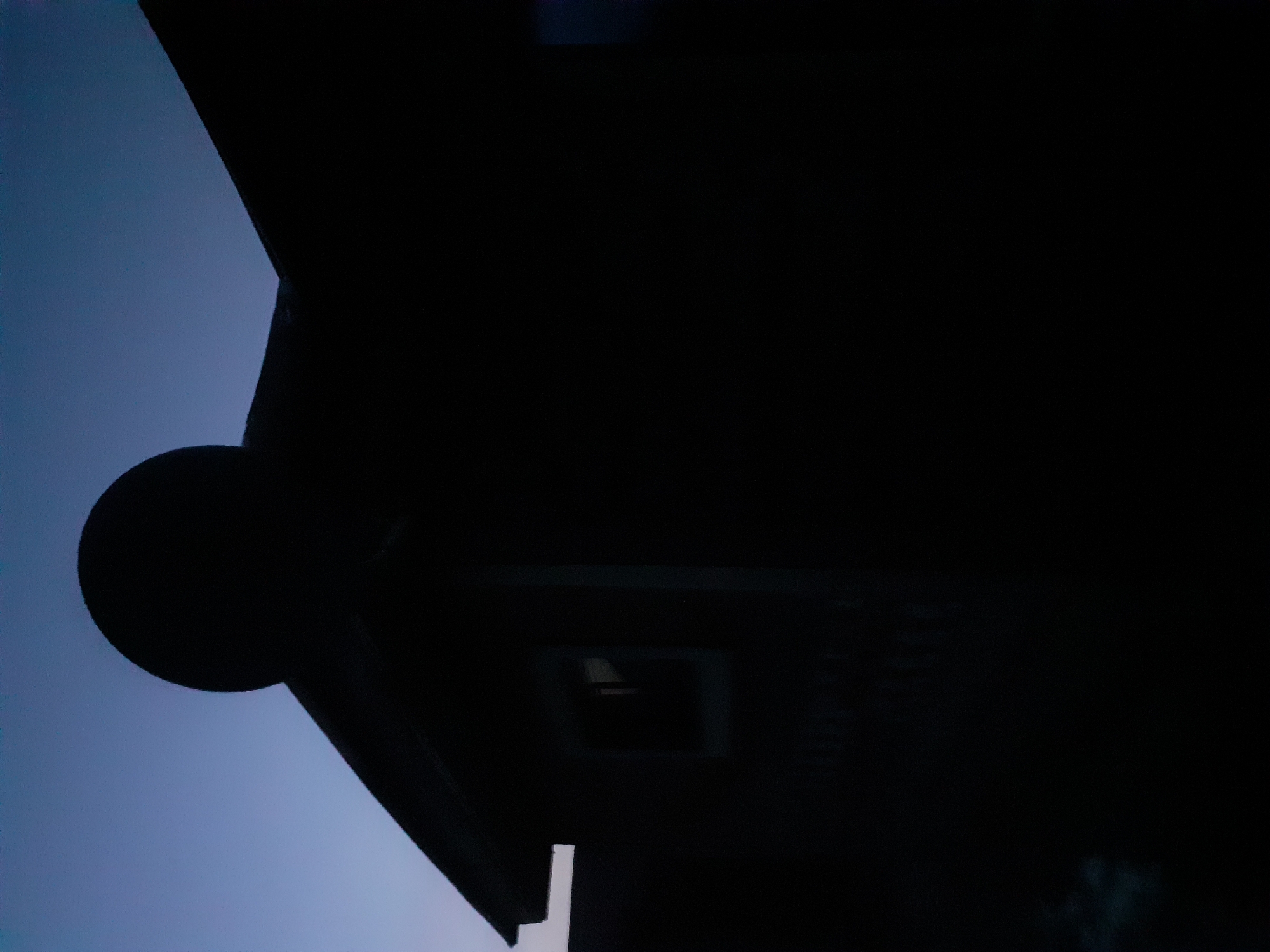 A silhouetted structure, possibly a small building or shed, is positioned against a twilight sky. A dark, circular object sits atop the structure's roof. A window is visible on the left side of the structure. The overall scene is dark, with the structure largely in silhouette against the fading light of the sky. The image does not provide enough information to determine its location in Jalingo, Nigeria.