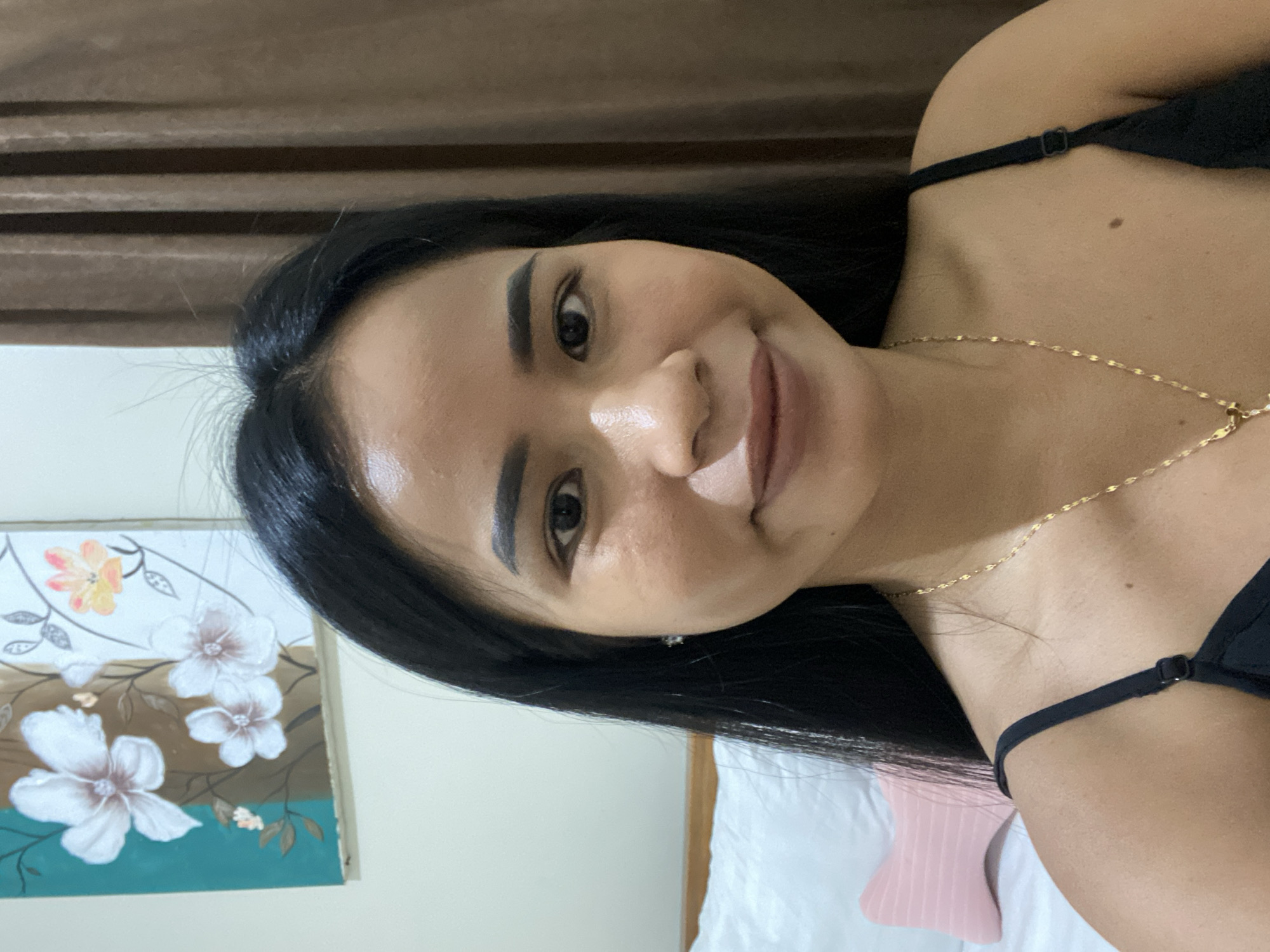 A woman is smiling at the camera. She has dark hair, brown eyes, and is wearing a gold necklace. She is wearing a black tank top with thin straps. The background includes a brown curtain, a painting with white flowers on a turquoise and brown background, a white pillow, and a pink object. The location is unknown, and there is no evidence to suggest the location is North Kuta, Indonesia.