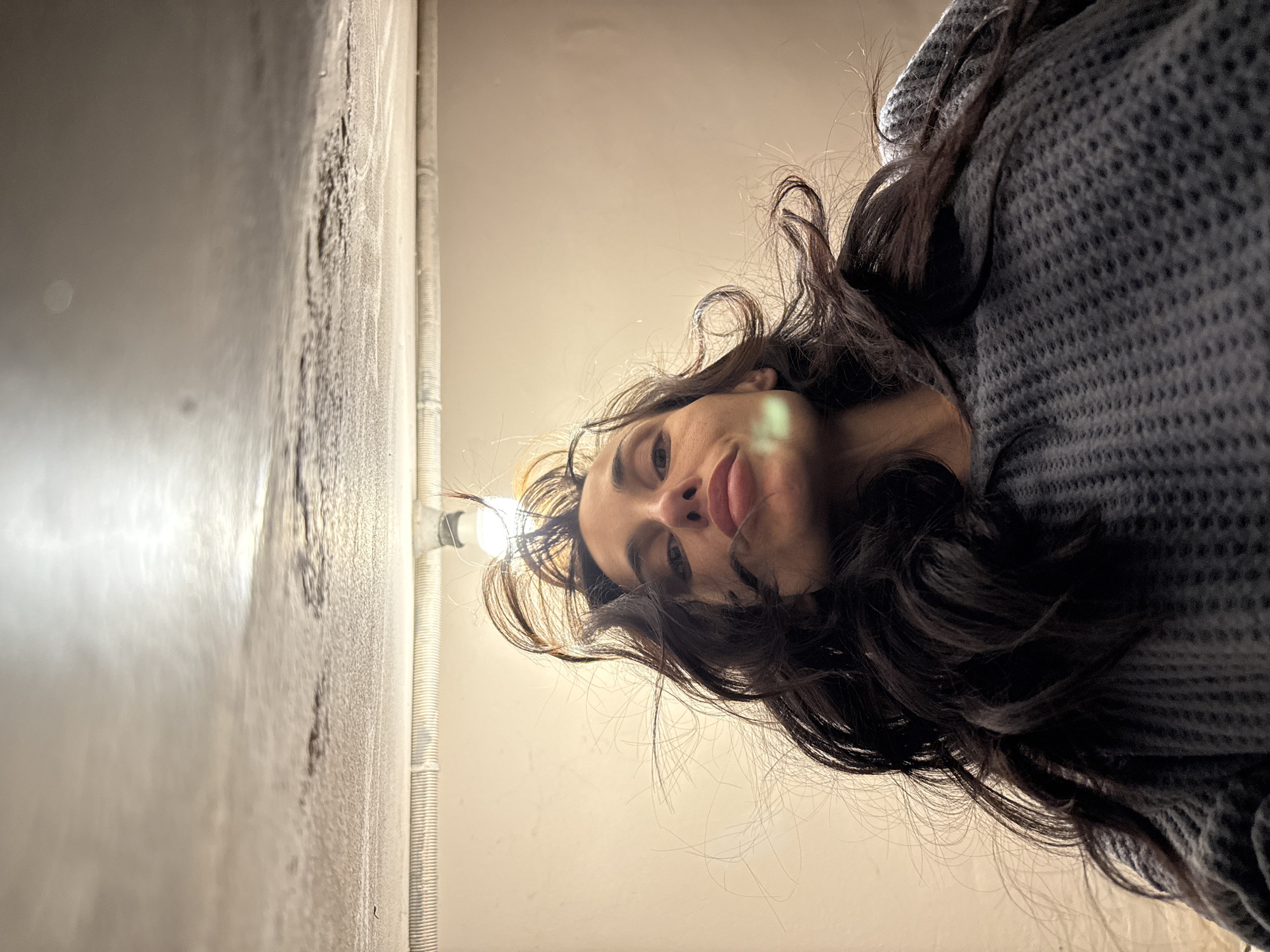 A low-angle shot depicts a person with long, dark, wavy hair looking down at the camera. They are wearing a gray knit sweater. Above the person is a pale yellow or beige ceiling. To the left is a textured wall with a light fixture. The location is not apparent from the image, and the claim that it is in Pleven, Bulgaria, cannot be substantiated.