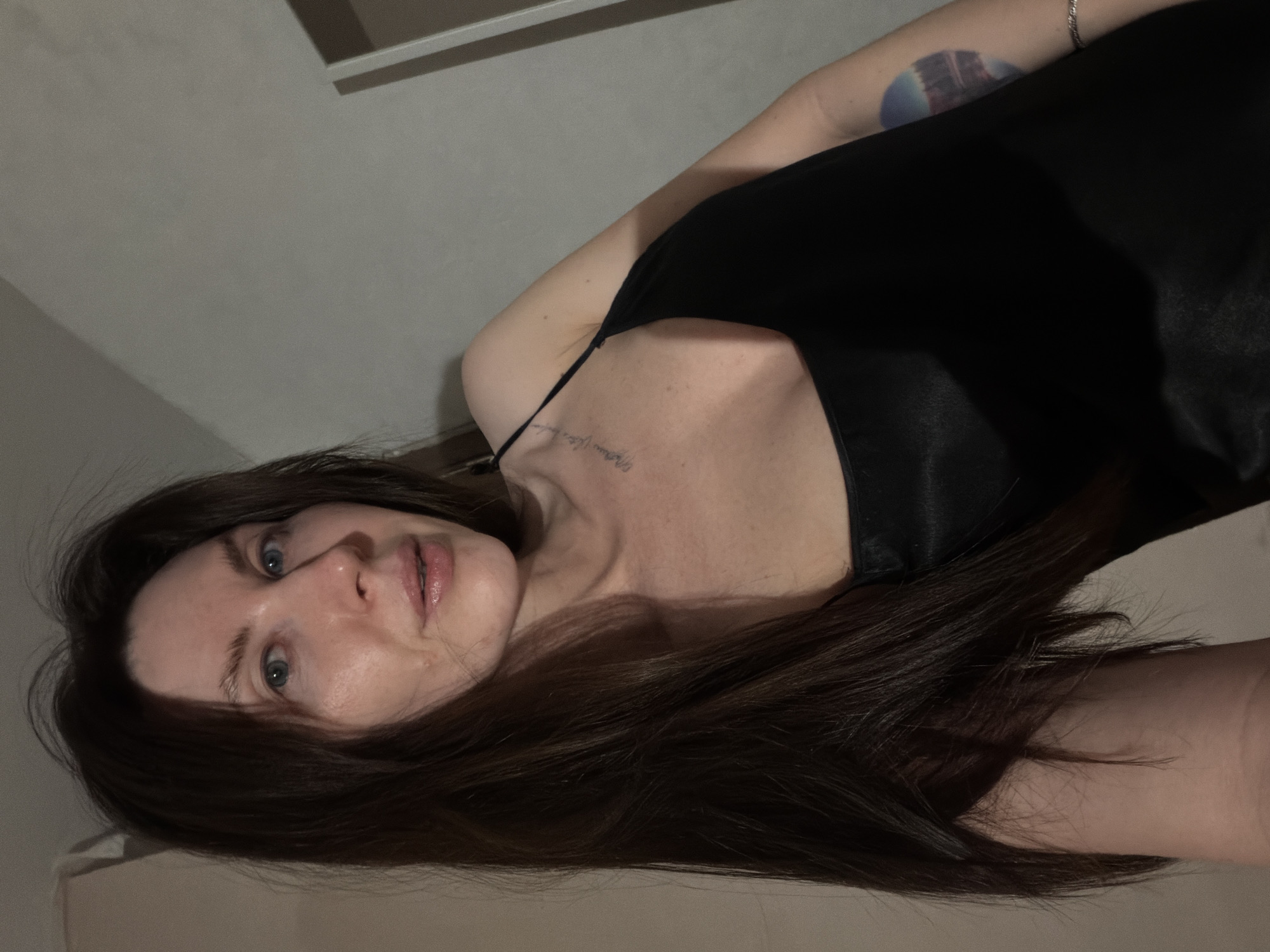 A woman is lying down, possibly on a bed. She has long brown hair and blue eyes. She is wearing a black top with thin straps. She has a tattoo on her collarbone and another one on her left arm. The background is a plain, neutral color, suggesting an indoor setting. There is no information available to confirm or deny if this image was taken in Perm, Russia.