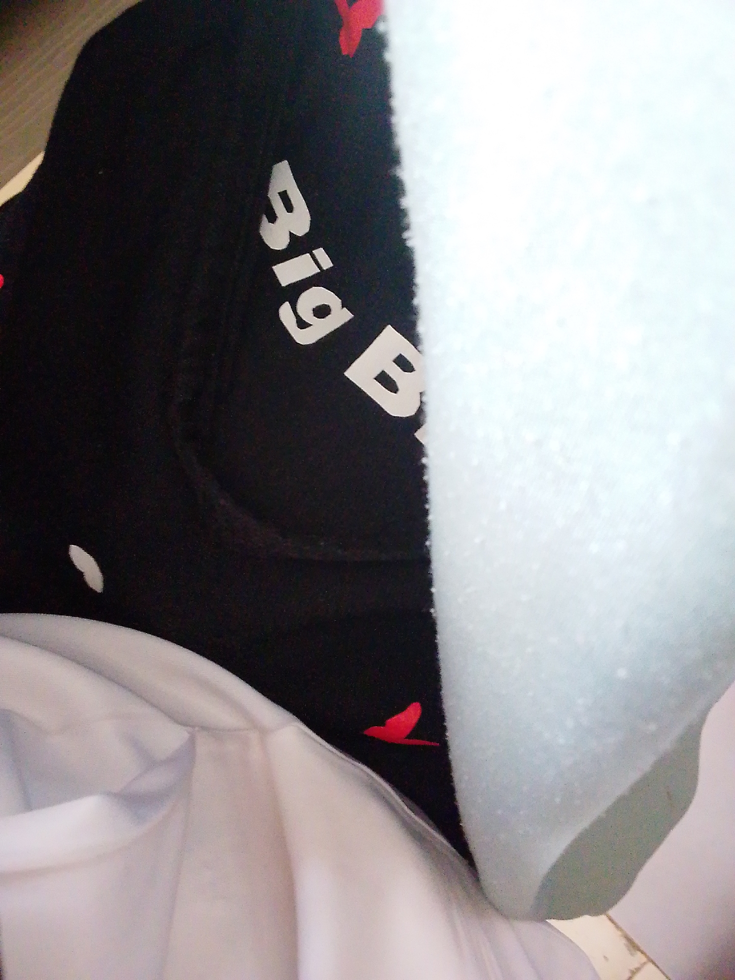 The media file shows a close-up of a black fabric with white text and red bird silhouettes. The text reads "Big B" in a stylized font. The fabric appears to be part of an article of clothing, possibly a hat or a shirt, with a white garment visible underneath. A pale blue, textured material is also prominently featured, partially obscuring the black fabric. The lighting suggests an indoor setting, with no clear indication of the time of day or weather. The scene is abstract and focuses on the textures and patterns of the materials. There are no people visible, and the activity or event taking place is not discernible from the image. The overall impression is that of a still life or product shot, highlighting specific design elements of the black fabric. The setting is not clearly defined, but the context provided suggests a location in Kuson, Nigeria.