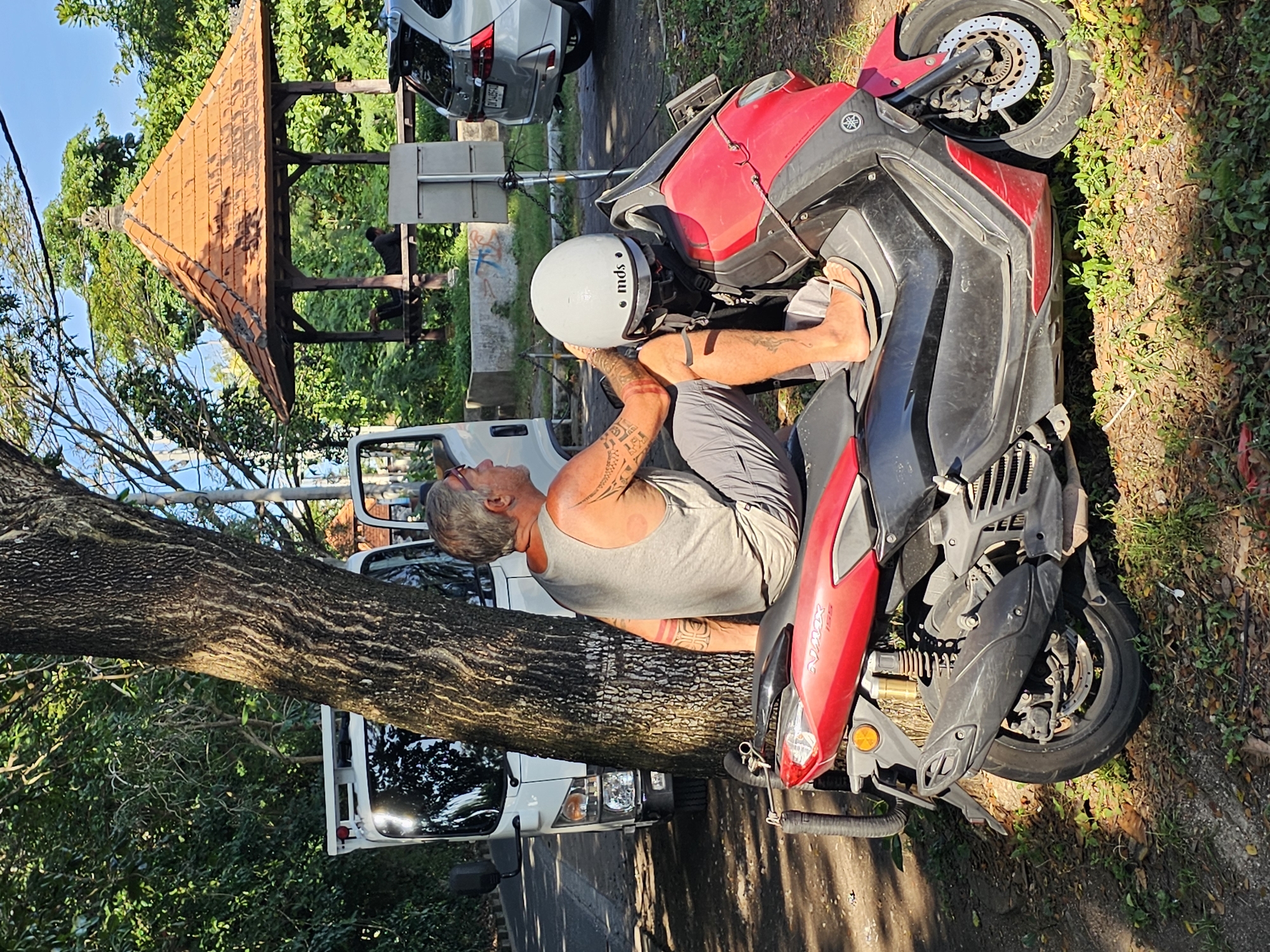A man with gray hair and tattoos on his arms sits on a red and black scooter parked on the side of a road in Denpasar, Indonesia. He wears a gray tank top and shorts. A white helmet rests on the scooter behind him. The scooter is positioned next to a large tree trunk. Behind the tree, a white van is parked. Further down the road, a gray car and a small pavilion with an orange roof are visible. The environment includes vegetation and a concrete curb separating the road from the greenery.
