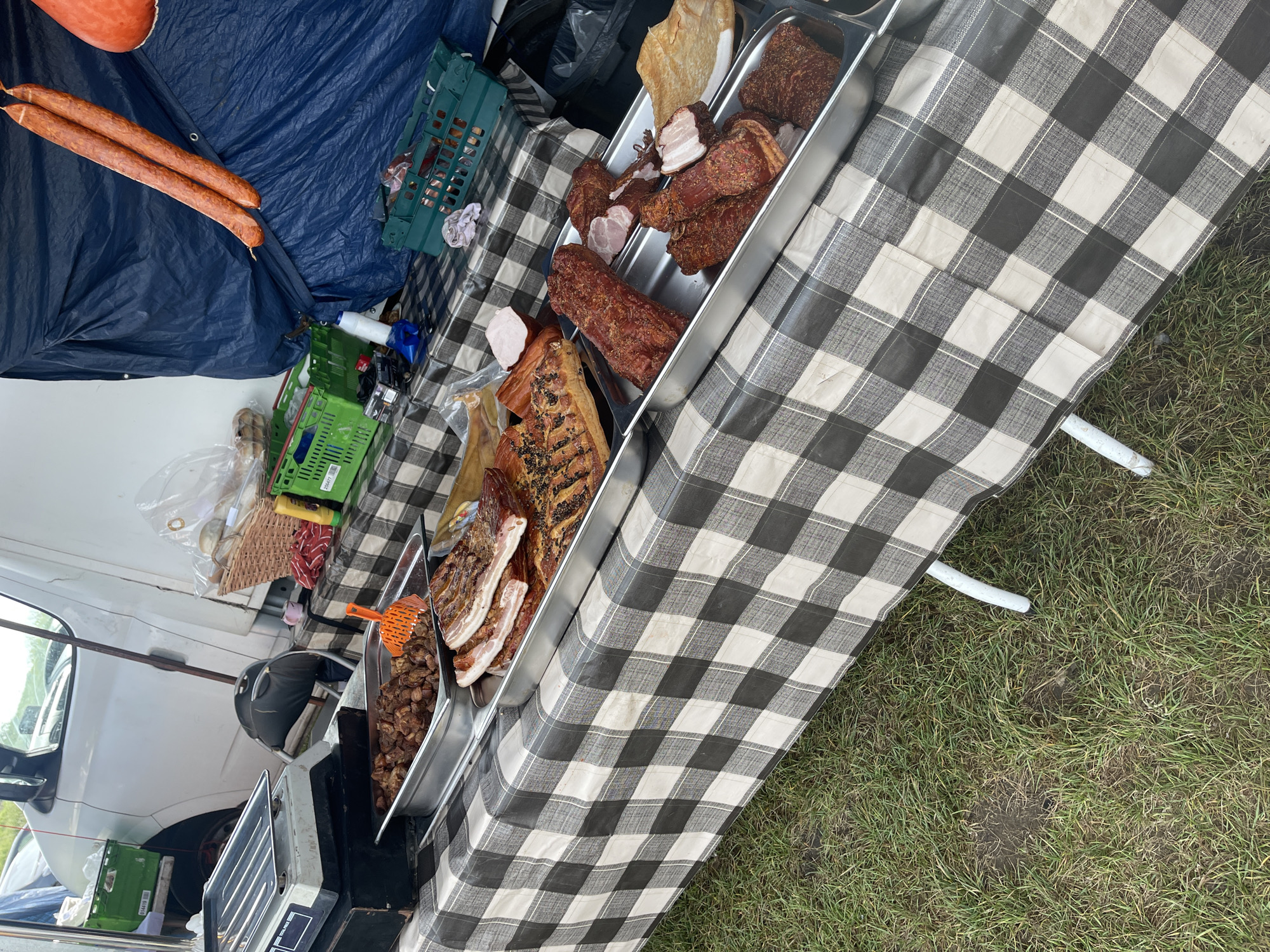 The image shows a market stall set up outdoors, likely in Sevenoaks, United Kingdom. The stall is covered with a black and white checkered tablecloth, displaying various meat products in silver trays. These include sausages, bacon, and what appears to be roasted pork belly. Behind the trays are crates, bottles, and packaging materials. A white van is visible in the background, indicating the stall is mobile. The stall's legs rest on grass.