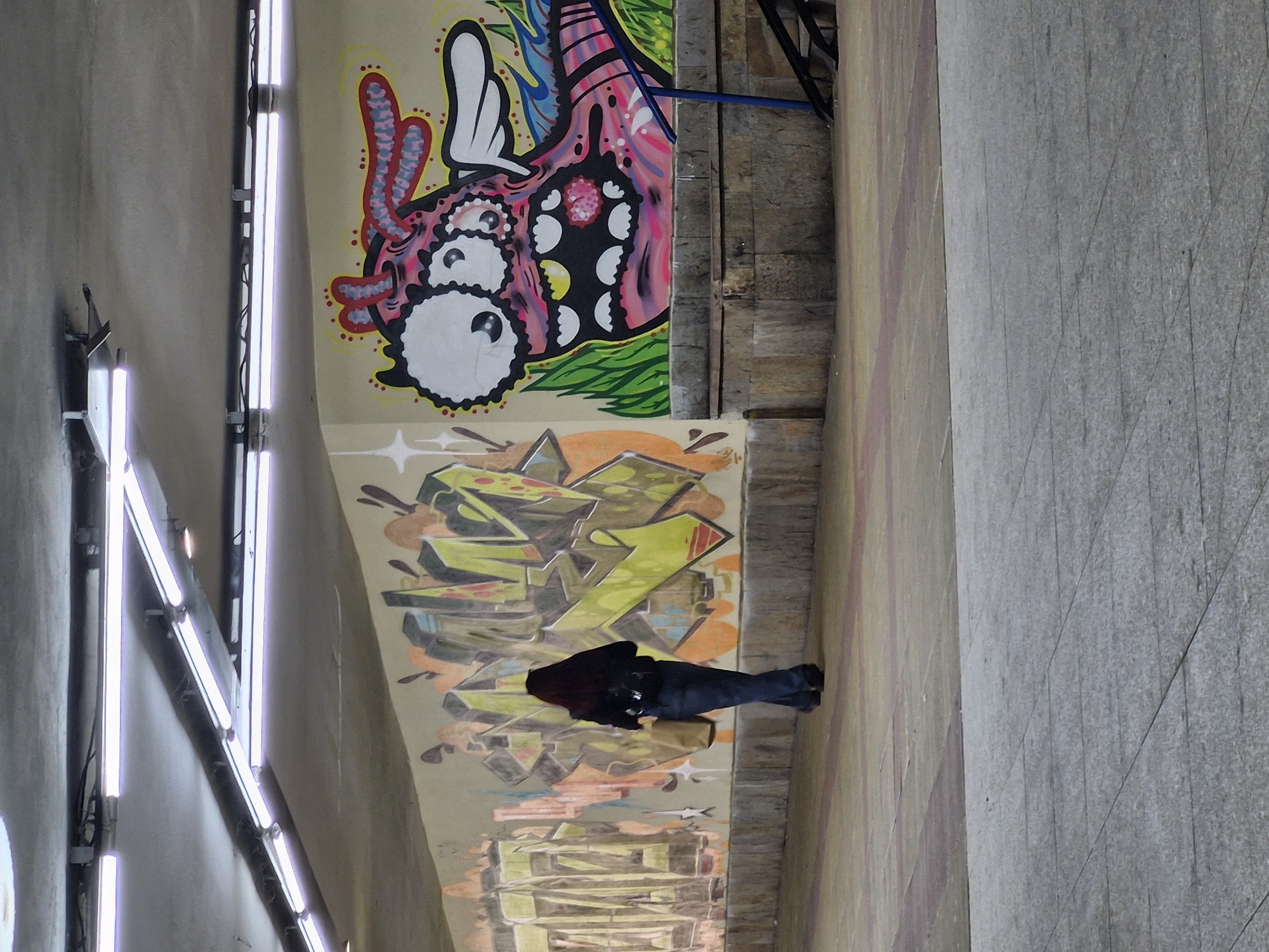 The image depicts an urban underpass or sheltered pathway in Sofia, Bulgaria, adorned with vibrant graffiti. In the foreground, a person is seen from behind, clad in a dark hooded top, blue jeans, and dark shoes. They appear to be leaning forward, looking down at something on the ground, with a light brown bag resting near their feet.
The main wall features two distinct styles of street art. The upper section showcases a large, colorful, cartoonish creature with a predominantly pink and black head, large white eyes with black pupils, and a wide mouth displaying white teeth. It has various red and blue tentacle-like appendages and is set against a background of green and blue abstract shapes on a tan wall. Below this, another layer of graffiti presents more angular, block-like designs in shades of yellow, green, orange, and brown, suggesting stylized letterforms.
To the left, a series of bright, vertical fluorescent light tubes illuminate the scene, mounted on a dark frame against a light-colored wall. The ground and an adjacent wall are constructed from light grey concrete, showing visible seams and texture. The artificial lighting suggests the scene is either indoors, an underpass, or captured during the evening or night. There is no clearly legible text visible in the graffiti or on the surrounding surfaces.