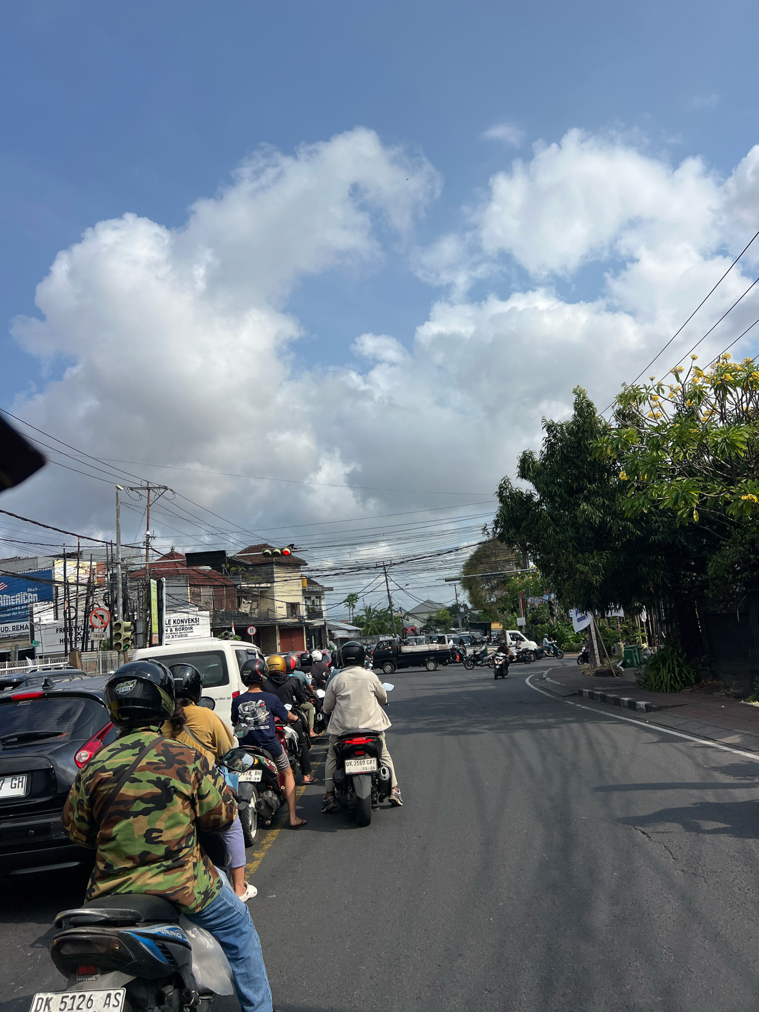 The image depicts a busy street scene in Denpasar, Indonesia, likely during daylight hours, with heavy traffic. A multitude of motorcycles and a few cars are present on the asphalt road, which appears to be a main thoroughfare. The sky is a clear blue with scattered white cumulus clouds. Along the sides of the road, buildings of varying architectural styles are visible, some appearing to be commercial establishments with signage, while others seem to be residential structures. Utility poles with a dense network of electrical wires crisscross overhead. Lush green trees and foliage line parts of the street, adding a natural element to the urban environment. Several individuals are seen riding motorcycles, most wearing helmets for safety. The overall impression is one of a vibrant and active urban setting characterized by significant vehicular traffic and the daily rhythm of city life.