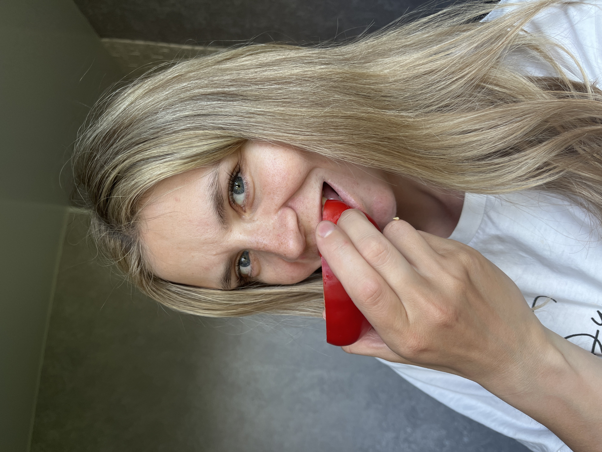 A Caucasian woman with long blonde hair and blue eyes is holding a red object to her mouth. She is wearing a white t-shirt and a gold ring on her right hand. The background is a gray wall. The location is unknown, and there is no information to suggest it is Chaikovsky, Russia.