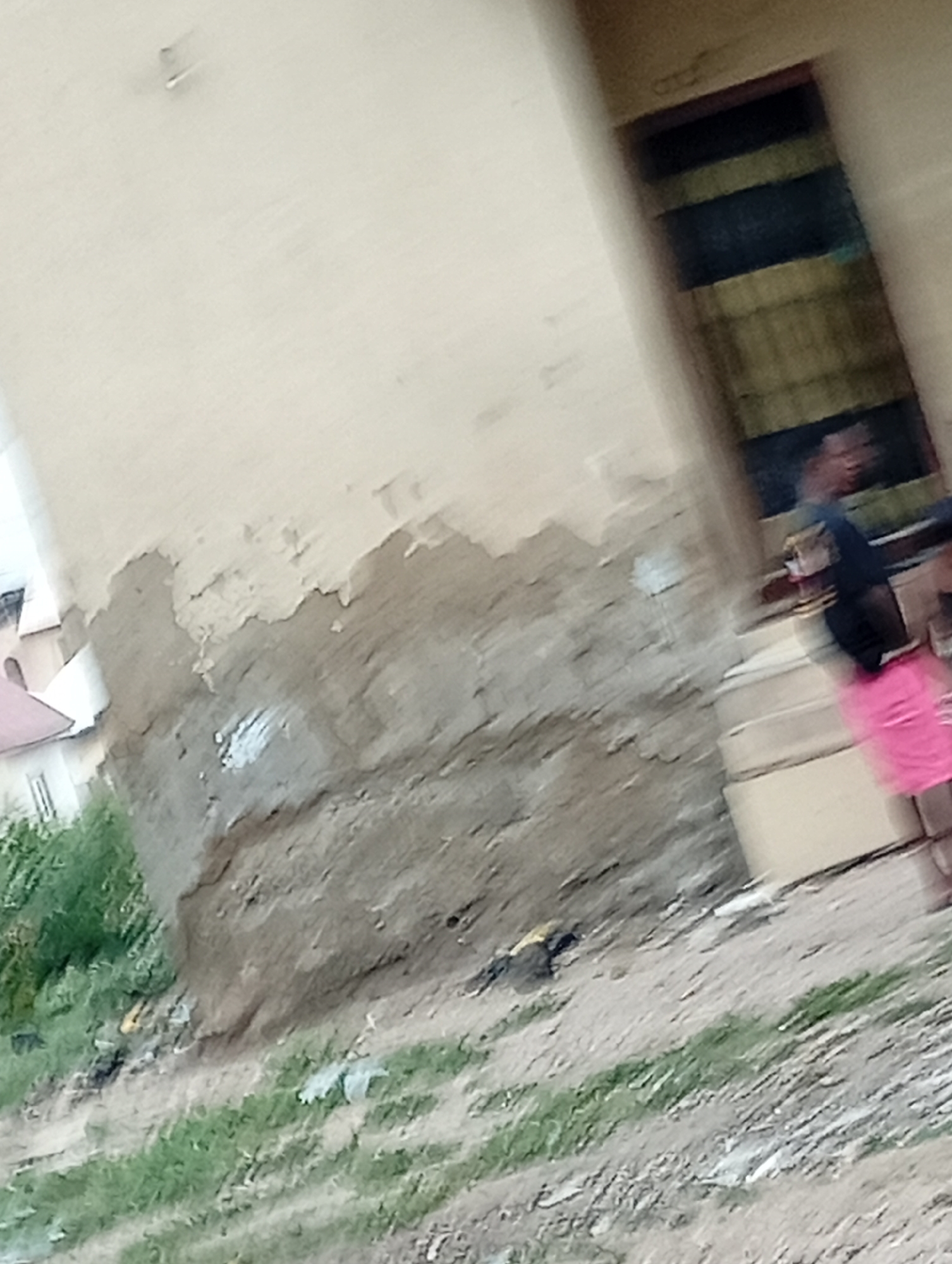 The image depicts a building exterior in what is described to be Adankolo, Nigeria. The building is of concrete construction with signs of age and disrepair visible, specifically, peeling paint and weathered surfaces. A portion of what appears to be the building's foundation is exposed near ground level. Vegetation and some debris are visible near the base of the building. A partially obscured individual wearing a dark shirt and pink shorts stands near an open doorway or entrance of the building. The image is somewhat blurry and taken at an angle.