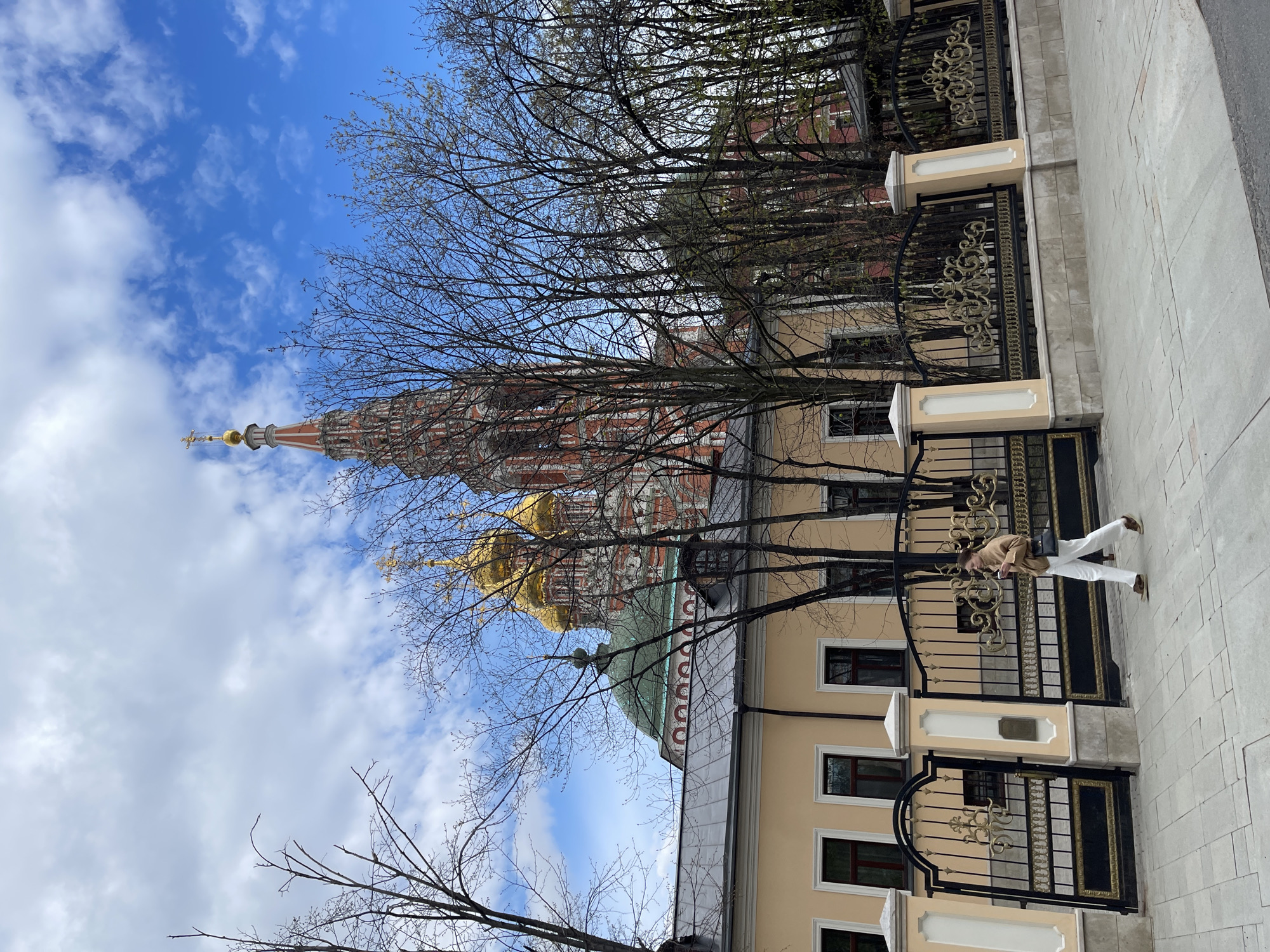 The image captures a scene in Moscow, Russia, featuring architectural details and a pedestrian. In the background, a building with multiple golden domes and a red and white tower rises, suggesting a cathedral or similar religious structure. A building with a light yellow facade stands in the foreground, partially obscured by bare trees.
A black and gold ornate fence runs along the side of the building, parallel to a light gray stone sidewalk. A person wearing a tan jacket and white pants is walking on the sidewalk next to the fence. The sky above is bright blue with scattered white clouds.