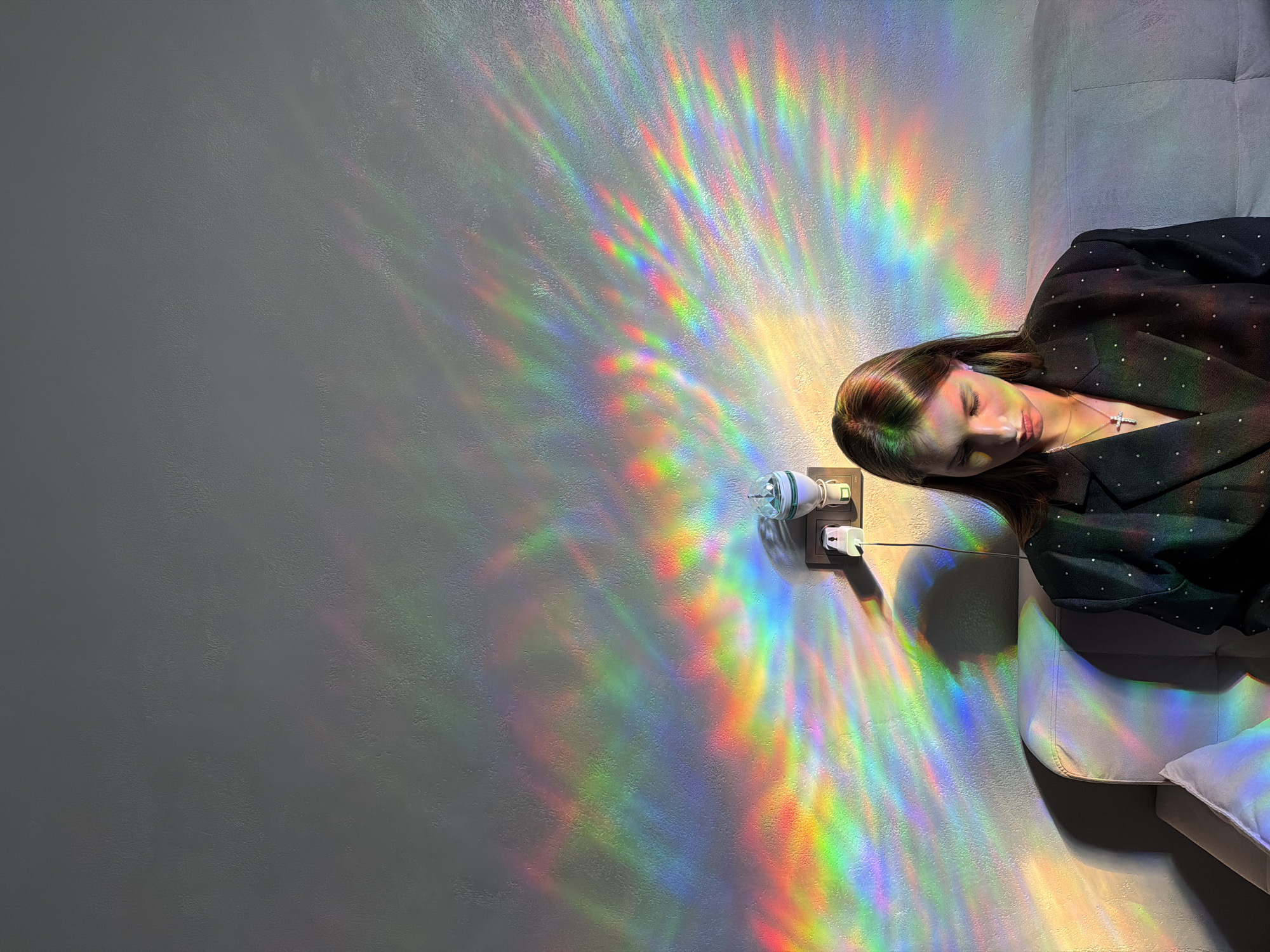 A woman is lying on a couch, her eyes closed, appearing to be asleep or resting. She is wearing a dark jacket adorned with small, scattered rhinestones and a silver necklace with a cross pendant. Her hair is dark brown and falls over her shoulder. The scene is illuminated by a light fixture plugged into a wall socket, which is projecting a vibrant rainbow of light patterns onto the textured gray wall and the couch. The light creates a captivating, abstract display of colors. Next to the woman, a white pillow is visible on the couch. The overall mood is serene and artistic, with the light effects adding a touch of magic to the quiet moment. The background suggests an indoor setting, possibly a home or a studio. There is no visible text in the image. The time of day and weather are not explicitly indicated, but the artificial lighting suggests it could be evening or a dimly lit interior.