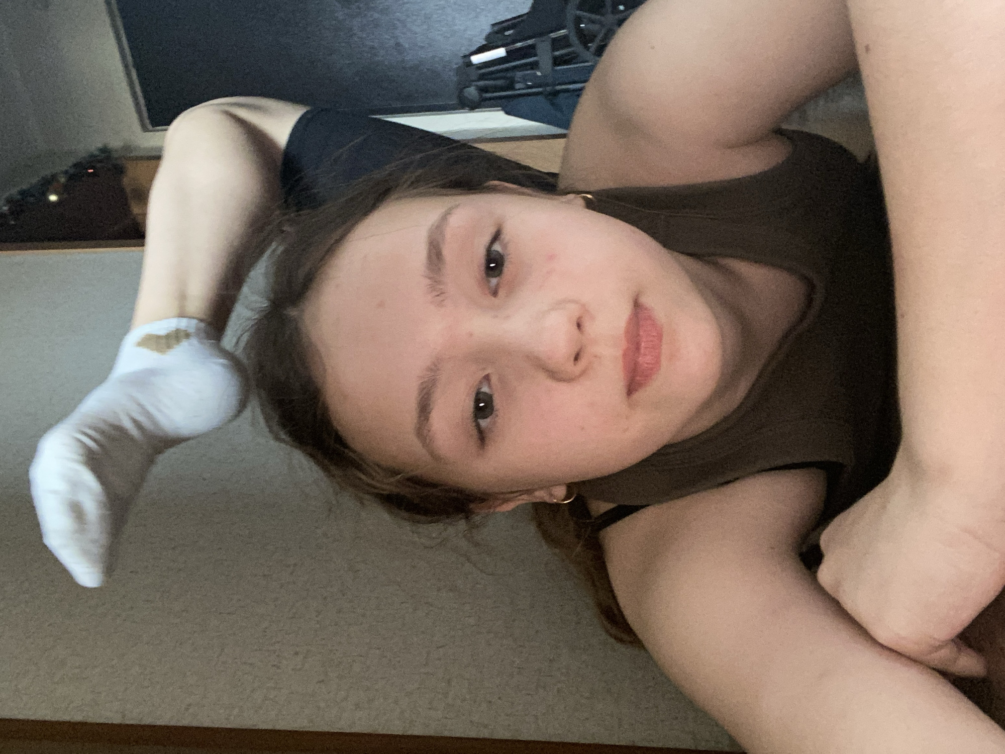 A young person lies prone on a carpeted floor, with their left leg arched over their head and the foot in a white sock. The person is wearing a brown tank top and dark shorts. Their face is visible, showing neutral expression. A metal folding object rests near the wall in the background.