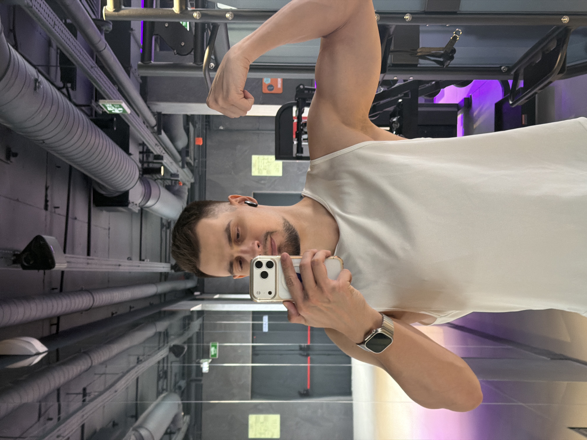 A man is taking a selfie in a mirrored gym. He is flexing his bicep while holding a smartphone. He is wearing a white tank top and a watch. The gym has weightlifting equipment and exposed pipes on the ceiling. The exact location within Krasnoyarsk, Russia, cannot be determined from the image.