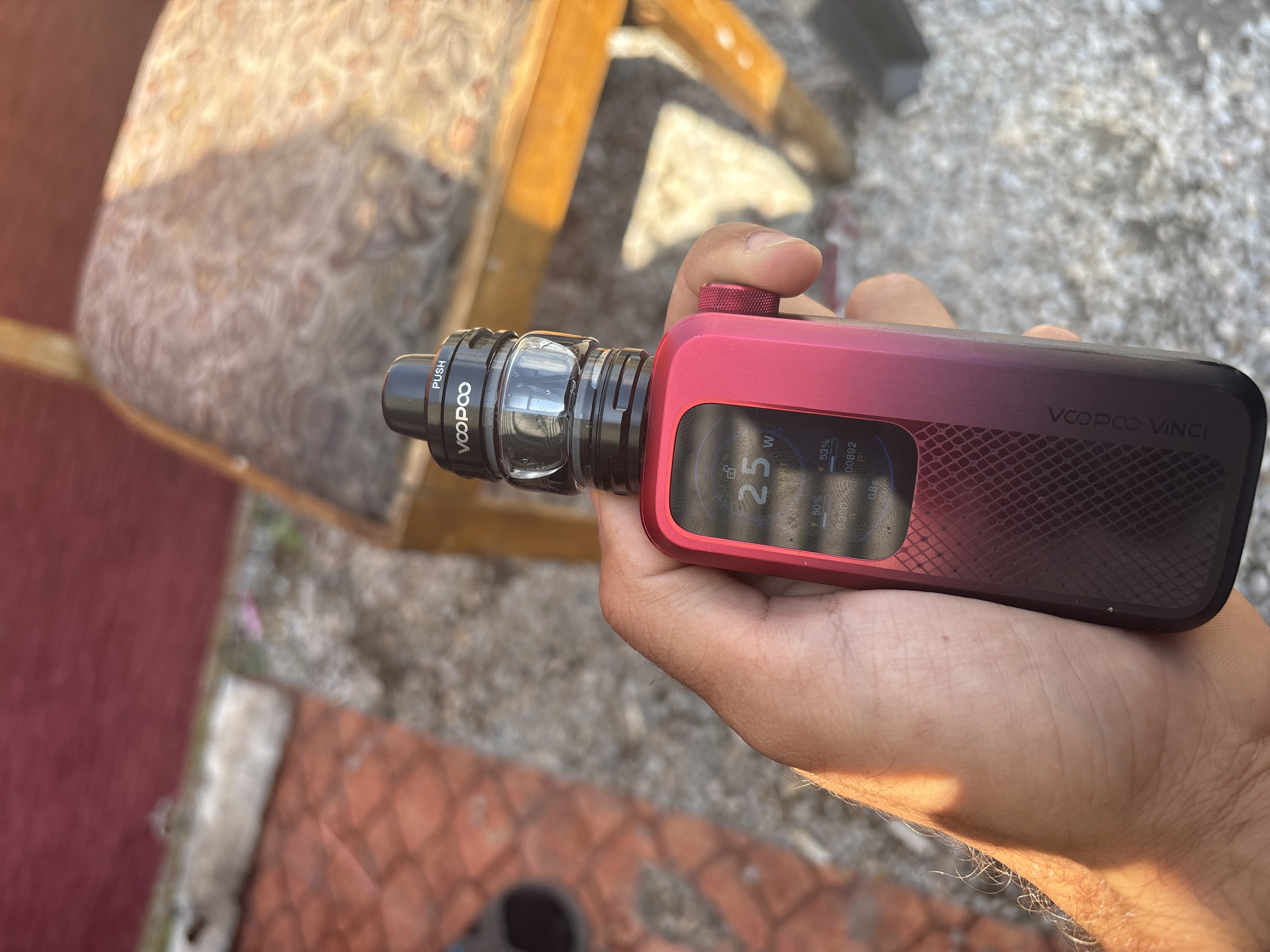 A close-up, outdoor shot captures a hand holding a red and black VOOPOO VINCI vape device, likely in Karadjovo, Bulgaria. The device features a clear tank with a visible e-liquid, and a black top section engraved with "PUSH" and "VOOPOO". The main body of the device displays "VOOPOO VINCI" on its side. Its screen is illuminated, showing various settings: "25 W", "50%", "53%", "0.30Ω", "0.8s", and "00892 P", along with the word "SMART".

The hand holding the vape is illuminated by bright sunlight, suggesting a clear, sunny day. The background is out of focus, revealing hints of an outdoor setting. Directly behind the vape, there's a blurred upholstered chair with a patterned fabric and wooden frame. Further back, the ground appears to be gravel or small stones, transitioning to a reddish paved or tiled area. The strong sunlight casts distinct highlights on the device and the hand, and potentially indicates mid-morning or afternoon.