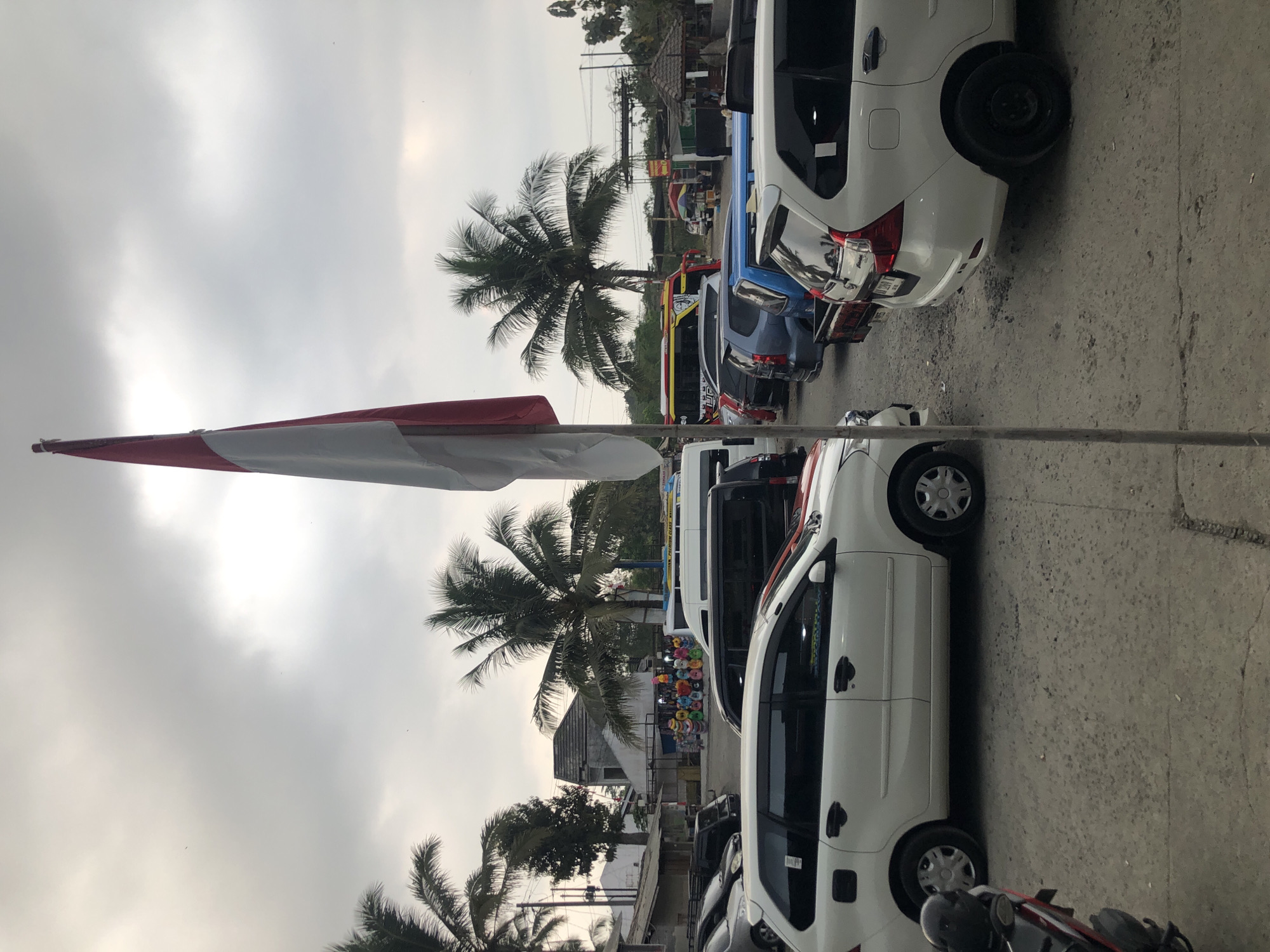 This image depicts a street scene in Borobudur, Indonesia, on a cloudy day. A prominent Indonesian flag stands tall in the foreground, its red and white colors stark against the overcast sky. The street is filled with parked vehicles, including white MPVs, a black van, and a colorful bus. Palm trees are visible in the background, suggesting a tropical environment. Stalls selling various items, possibly souvenirs or food, are set up along the roadside, with a cluster of colorful hats visible at one stall. The ground is a mixture of asphalt and dirt. The overall impression is that of a busy area, possibly near a tourist attraction or transportation hub.