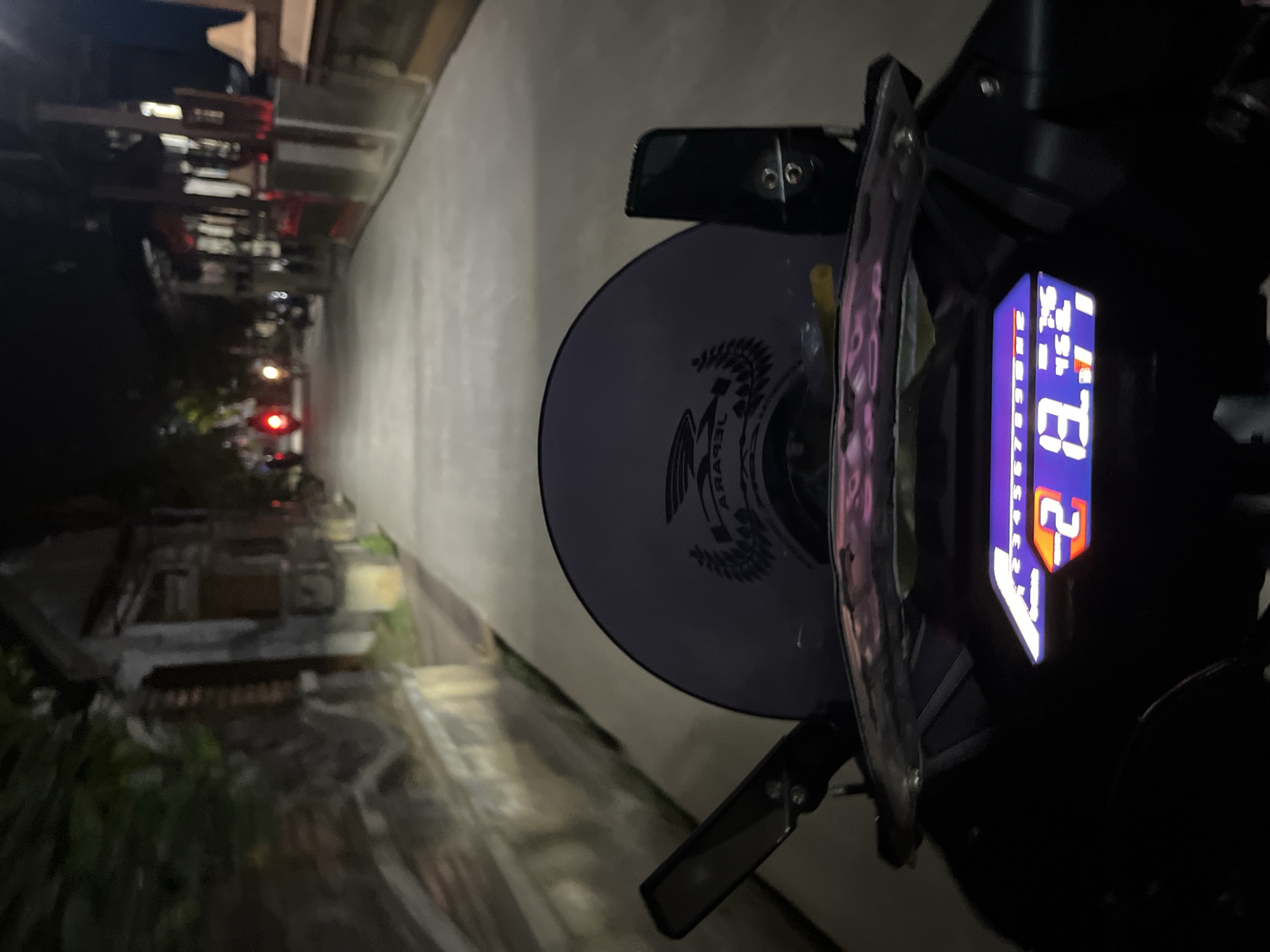 A first-person perspective shot from a motorcycle at night, shows a paved road lined with buildings on either side. A digital speedometer is visible in the foreground, displaying a speed of 20 km/h. A dark tinted windshield with a logo is in front of the rider. In the distance, other motorcycles with illuminated tail lights can be seen. The scene appears to be in Karangrandu, Indonesia, given the visual cues of the buildings and the road.