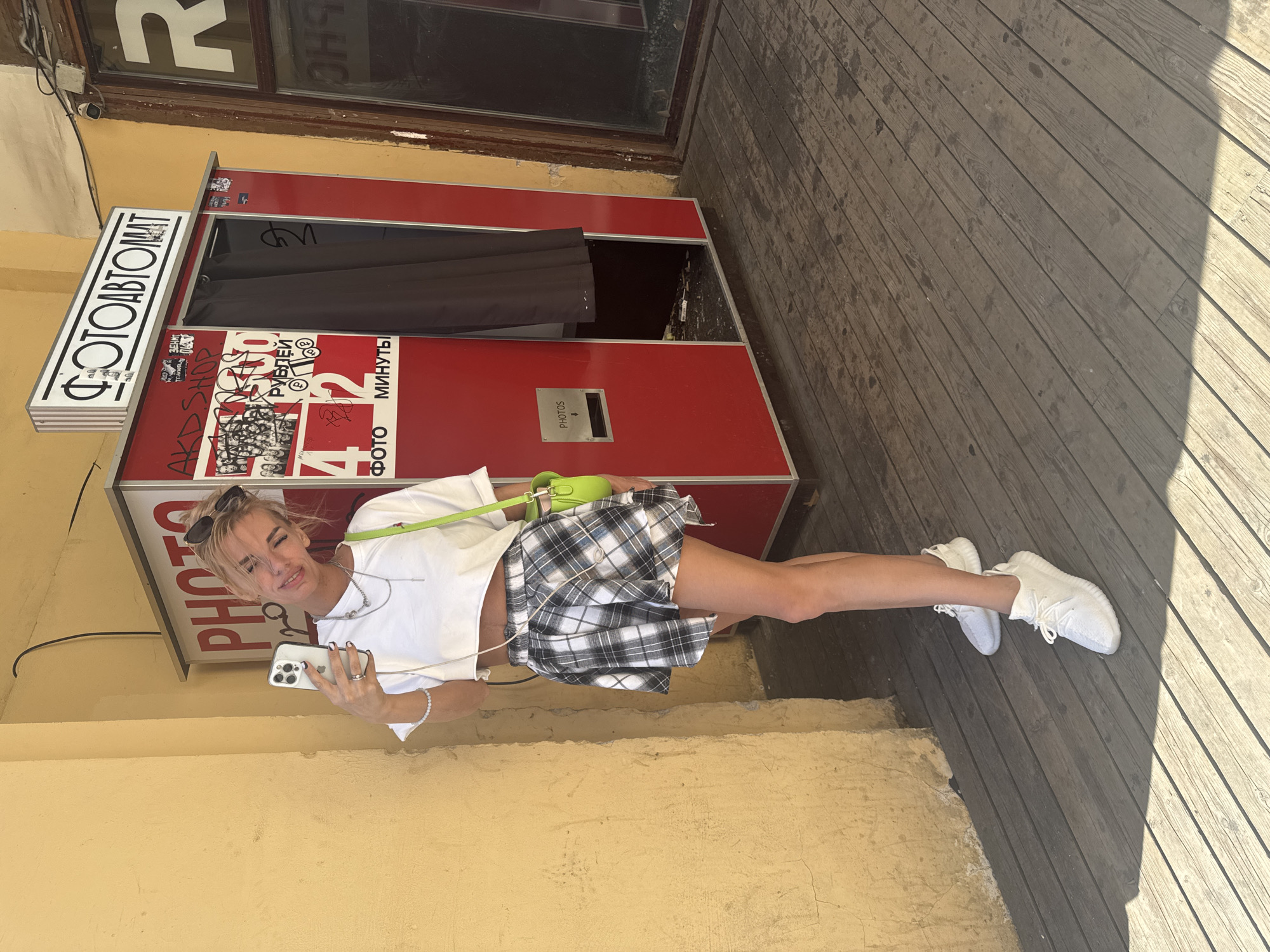 A young Caucasian woman with blonde hair and sunglasses is taking a selfie with her smartphone in front of a red photo booth machine. She is wearing a white crop top, a black and white plaid skirt, and white sneakers. The photo booth has Russian text that translates to "Photoautomat." It is located on a wooden boardwalk next to a yellow building. The photo booth has a slot for photos and appears to be in a public outdoor space in Saint Petersburg, Russia.