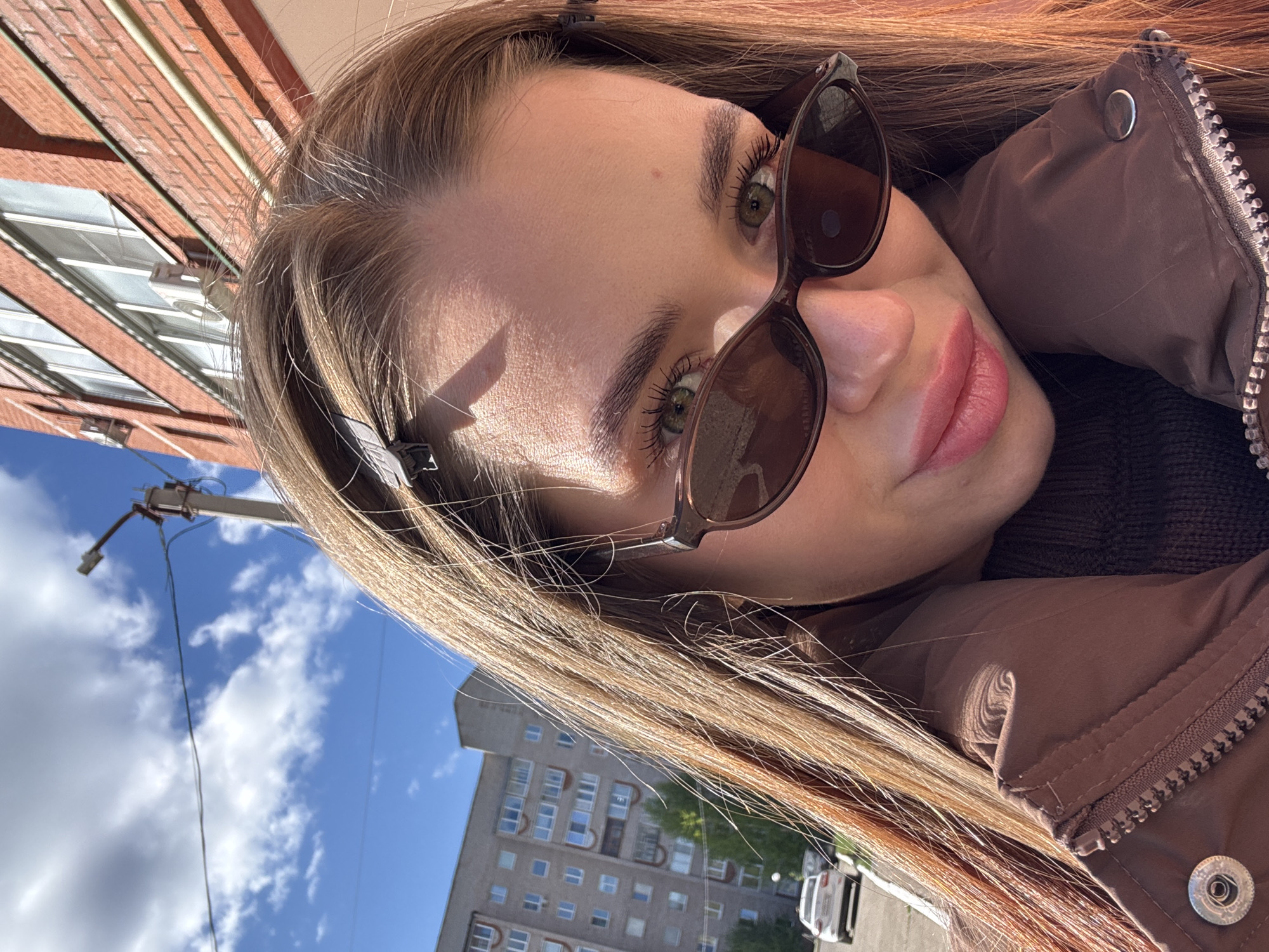 The image shows a selfie of a young woman with light brown hair, wearing brown sunglasses and a brown jacket. She is looking directly at the camera with green eyes and pink lips. The background includes a brick building with windows, a blue sky with white clouds, an apartment building, a utility pole, and a white car. The location is Chaikovsky, Russia.