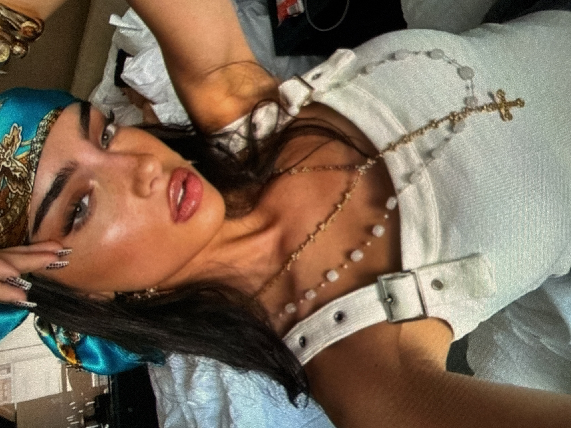 A woman is lying down, wearing a patterned turquoise headscarf, a white top with buckles, and a rosary necklace. Her nails are decorated. She is indoors on a white surface. The city is unknown.