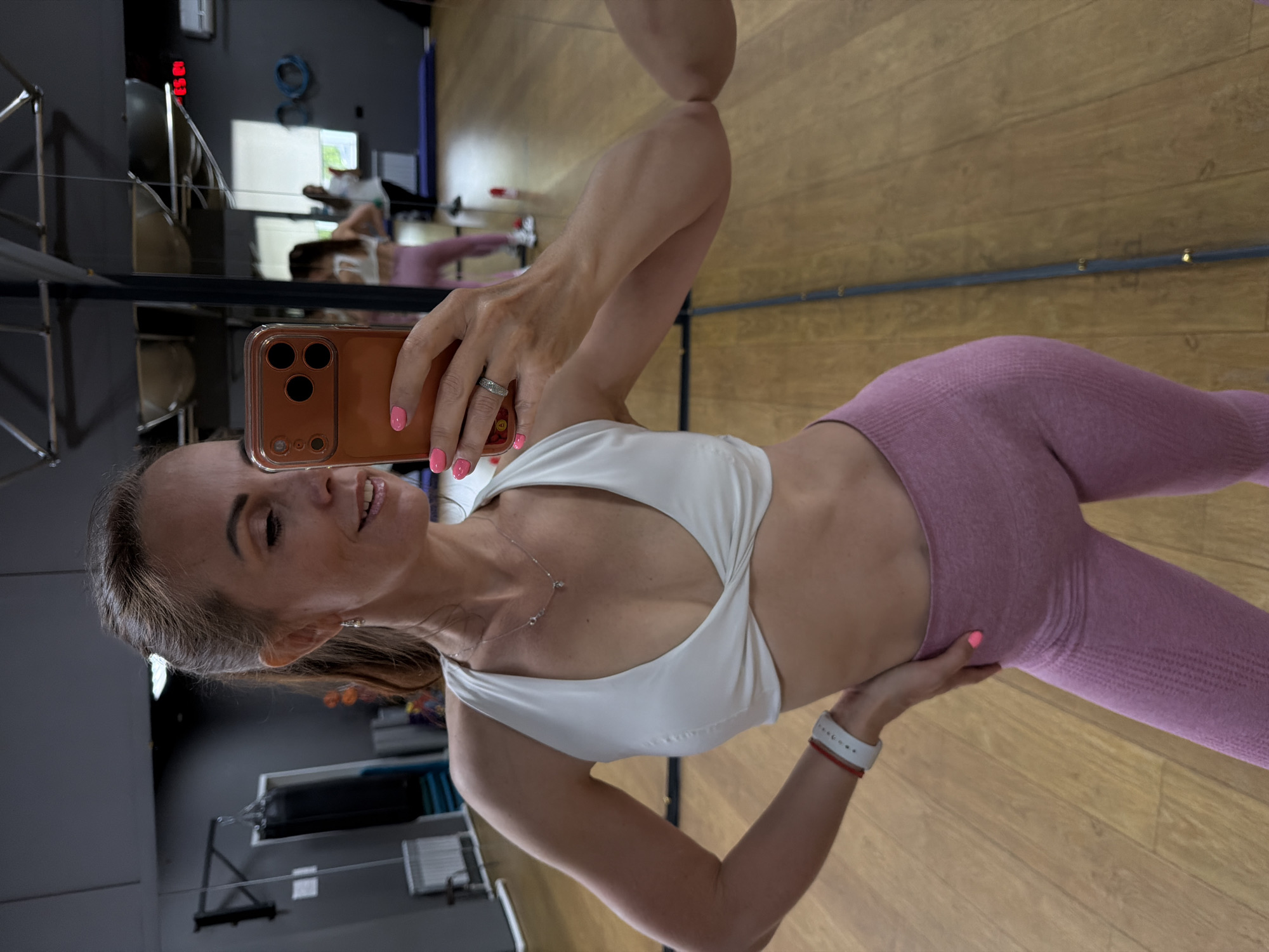 A woman with blonde hair tied in a ponytail, wearing a white sports bra and pink leggings, takes a mirror selfie in a gym. She is holding an orange smartphone in her right hand and has her left hand on her hip. Her nails are painted pink. In the reflection, other people can be seen in the gym. The gym has a wooden floor and gray walls with various exercise equipment. The overall scene suggests a fitness-oriented environment.