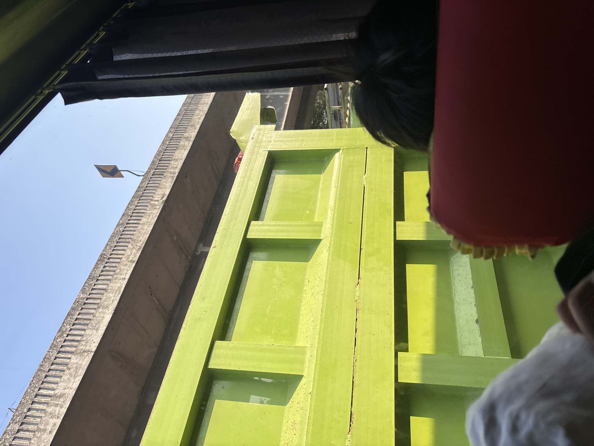 In Karawang, Indonesia, a bright green truck bed is prominently displayed in the foreground, filling the lower half of the frame. The truck's side features a sturdy, reinforced design with visible horizontal beams and vertical supports, suggesting a heavy-duty vehicle. Above the truck, a concrete overpass is visible, its underside showing a textured surface. A yellow directional arrow sign pointing left is affixed to the edge of the overpass. The sky above is a clear, pale blue. On the right side of the image, dark gray curtains are partially drawn. In the immediate foreground, a person's dark hair and the back of a red headrest of a seat are visible, indicating the photo was taken from inside a vehicle looking out. The lighting suggests it is daytime, with sunlight casting shadows and highlighting the textures of the truck and overpass.