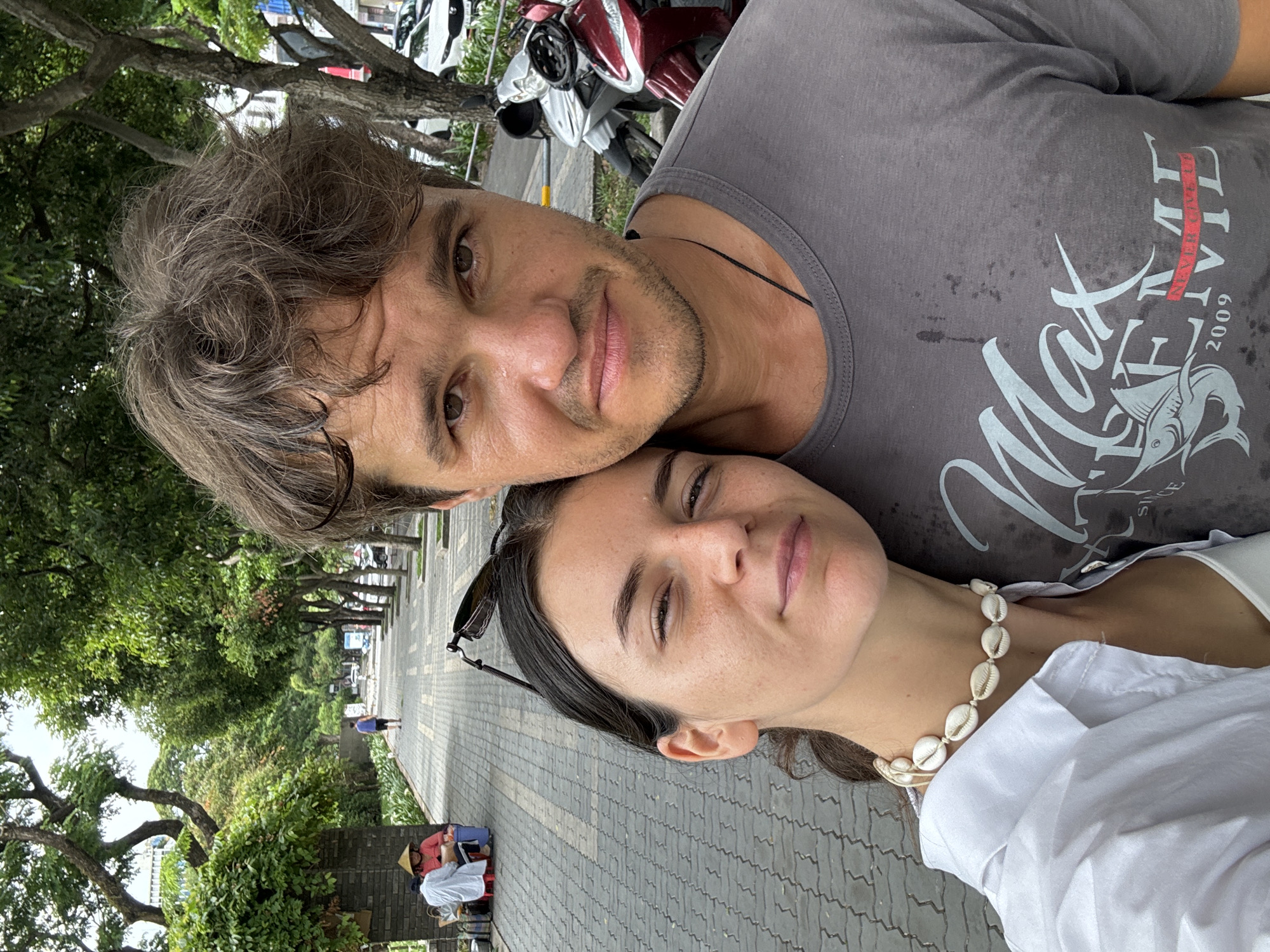 Two people are in a close-up selfie outdoors. A man with curly brown hair, a dark gray t-shirt with a logo, and stubble is positioned above a woman. The woman has dark hair pulled back, sunglasses perched on her head, a white shirt, and a seashell necklace. They are on a brick walkway. In the background is a street lined with trees, parked scooters, and a distant pedestrian. A small group sits on the side of the walkway. The location appears to be Thủ Đức, Vietnam, given the streetscape and vegetation.