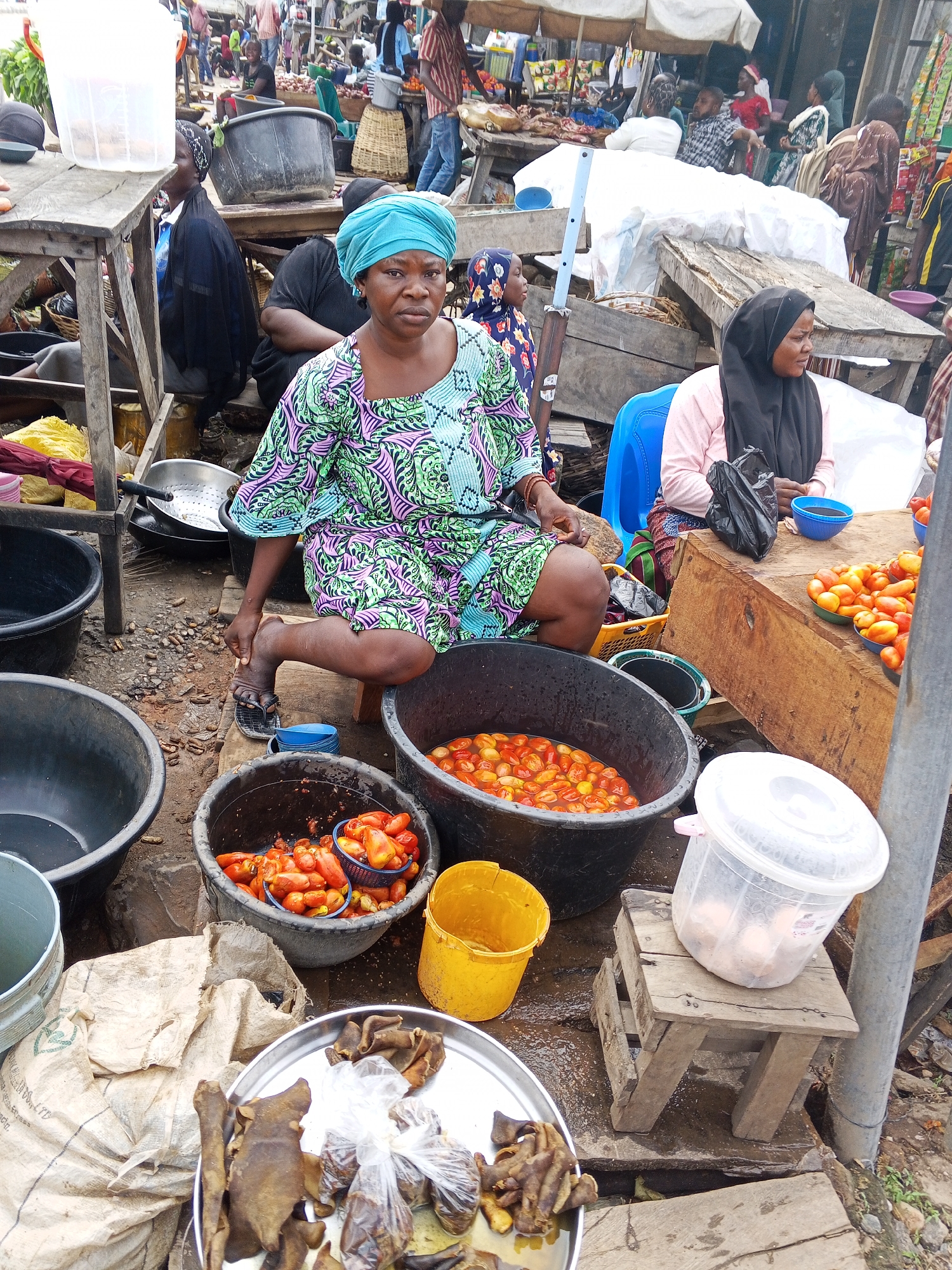 The image depicts a busy marketplace scene in Zango-ndaji, Nigeria. A woman wearing a vibrant, patterned dress and a blue headscarf is seated in the foreground, her right hand extended towards a black bucket filled with tomatoes. Beside her, to the right, another woman wearing a black hijab and a pink shirt is seated on a blue plastic chair, looking to her right. In front of the first woman are several large black buckets, one containing tomatoes, and a metal tray with what appears to be dried meat and packaged goods. To the right of the women, a wooden stall displays more tomatoes in bowls, and a blue bowl contains a dark liquid. In the background, other market-goers are visible, along with various stalls and produce, suggesting a lively commercial atmosphere. The ground is wet and appears to be dirt or mud, with some debris scattered around. The overall environment is characteristic of an outdoor African market.