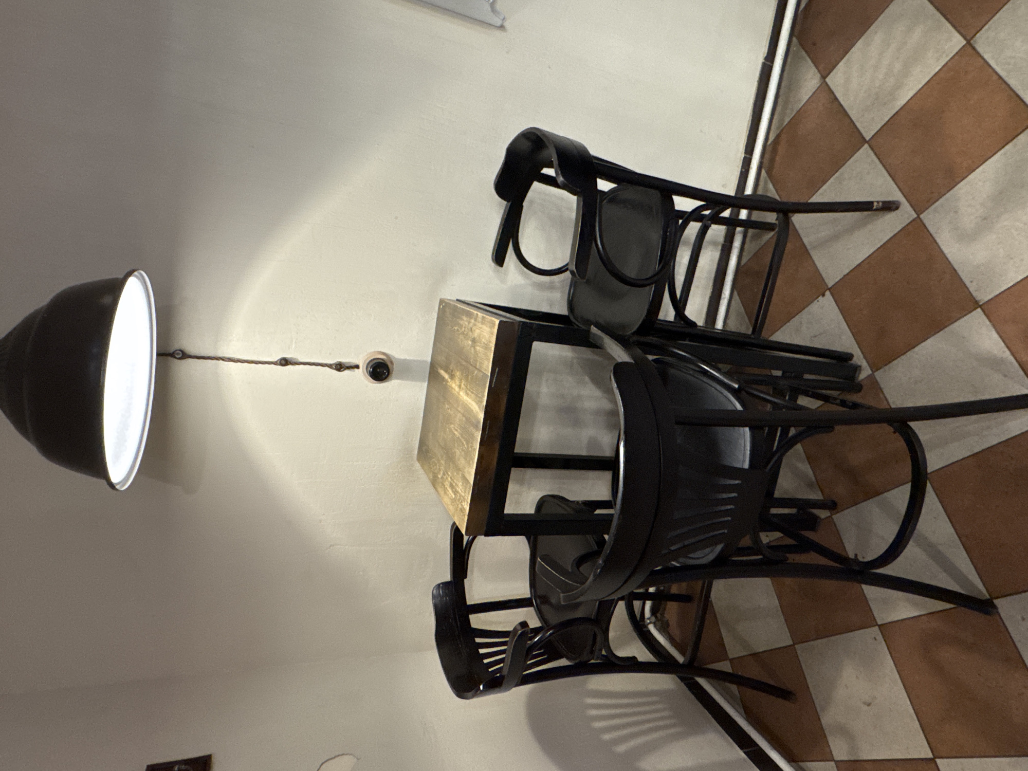 Here is a detailed description of the image:
The image shows a high-angle, slightly rotated shot of a cafe or dining area. A light fixture hangs from the ceiling, illuminating a small wooden table surrounded by three black chairs. The floor is tiled with alternating squares of brown and white. The scene appears indoors, with plain walls and a minimalist aesthetic. The photograph seems to focus on the furniture arrangement, suggesting a setup in a public establishment like a cafe. There are no people visible in the shot, and the overall atmosphere is quiet and still.