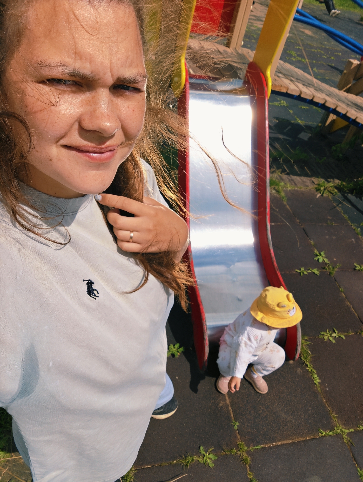 In this high-angle, candid selfie taken outdoors, a woman with long, light brown hair frames the left side of the image. She is wearing a light blue t-shirt with a black polo player logo. Her face is partially obscured by her hair, but she appears to be smiling slightly, with a relaxed expression. A simple gold band is visible on her left ring finger.
To the right of the woman, a young child is visible, facing away from the camera and squatting at the bottom of a red and yellow playground slide. The child is wearing a white outfit with a subtle pattern and a bright yellow hat adorned with a cartoon character. The child's head is tilted downwards, suggesting they are focused on something on the ground or about to slide.
The scene appears to be a playground, with the shiny metal surface of the slide reflecting the sunlight and the blue sky. A wooden bridge structure is visible behind the slide, part of the playground equipment. The ground is a dark, textured surface, likely rubberized playground material, with patches of green grass and weeds growing in the cracks. The lighting suggests it is a sunny day, likely daytime, with the sun casting bright highlights and shadows. The overall mood is casual and playful, capturing a moment between a woman and a child at a playground in Chaikovsky, Russia. There is no visible text in the image.