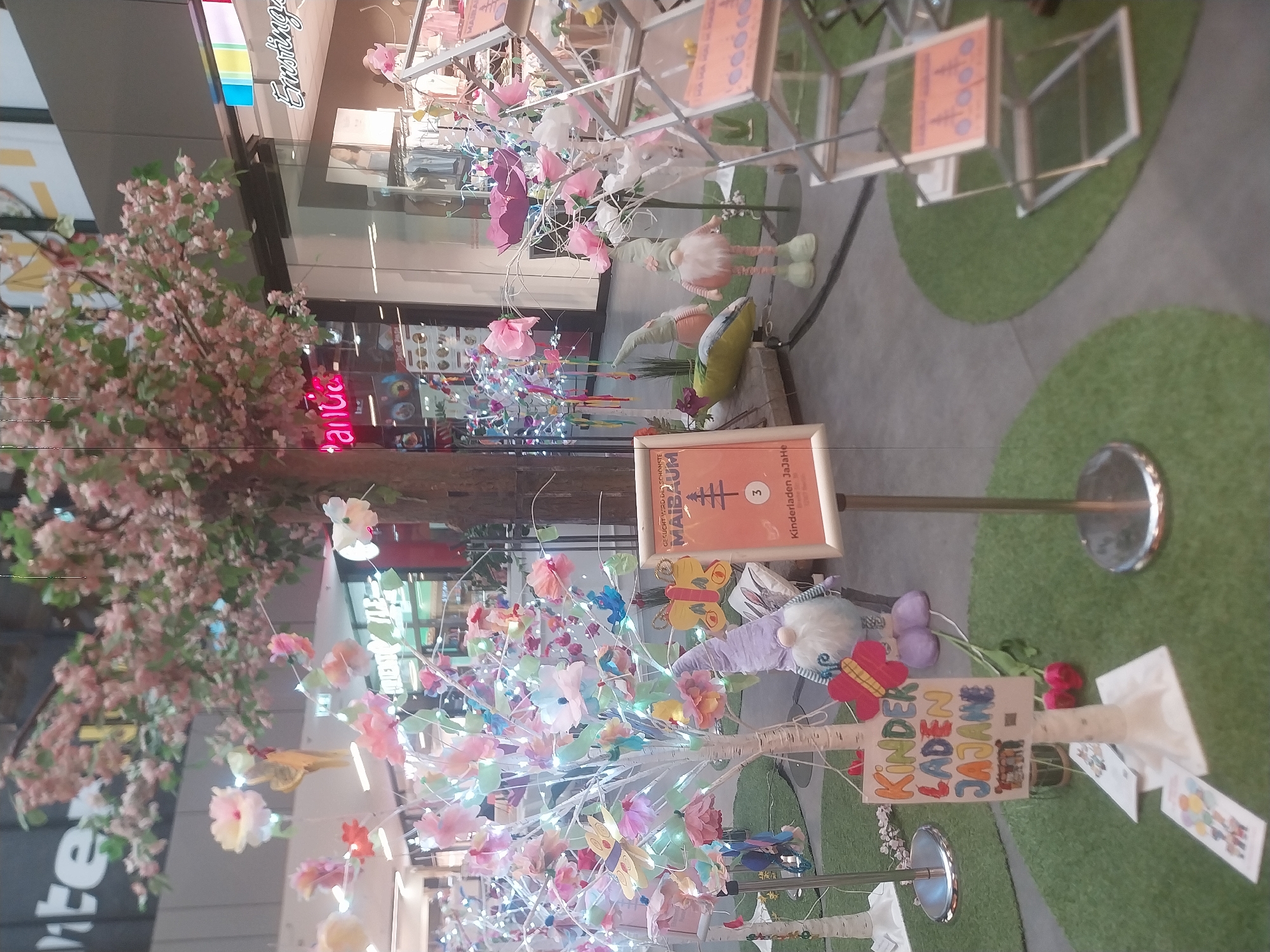 The image shows an indoor shopping area, likely a mall, decorated with artificial trees covered in paper flowers and fairy lights. The trees are set up on small circular patches of artificial grass. Signs are present with text including "Kinderladen JaJaHe" and "Kinder Laden JAJAME," suggesting a children's store. One sign reads "GESUCHT WIRD DER SCHÖNSTE MAIBAUM" followed by a tree symbol and the number 3. There are also figures of gnomes. The store is likely in the Emsringe shopping center in Berlin, Germany.