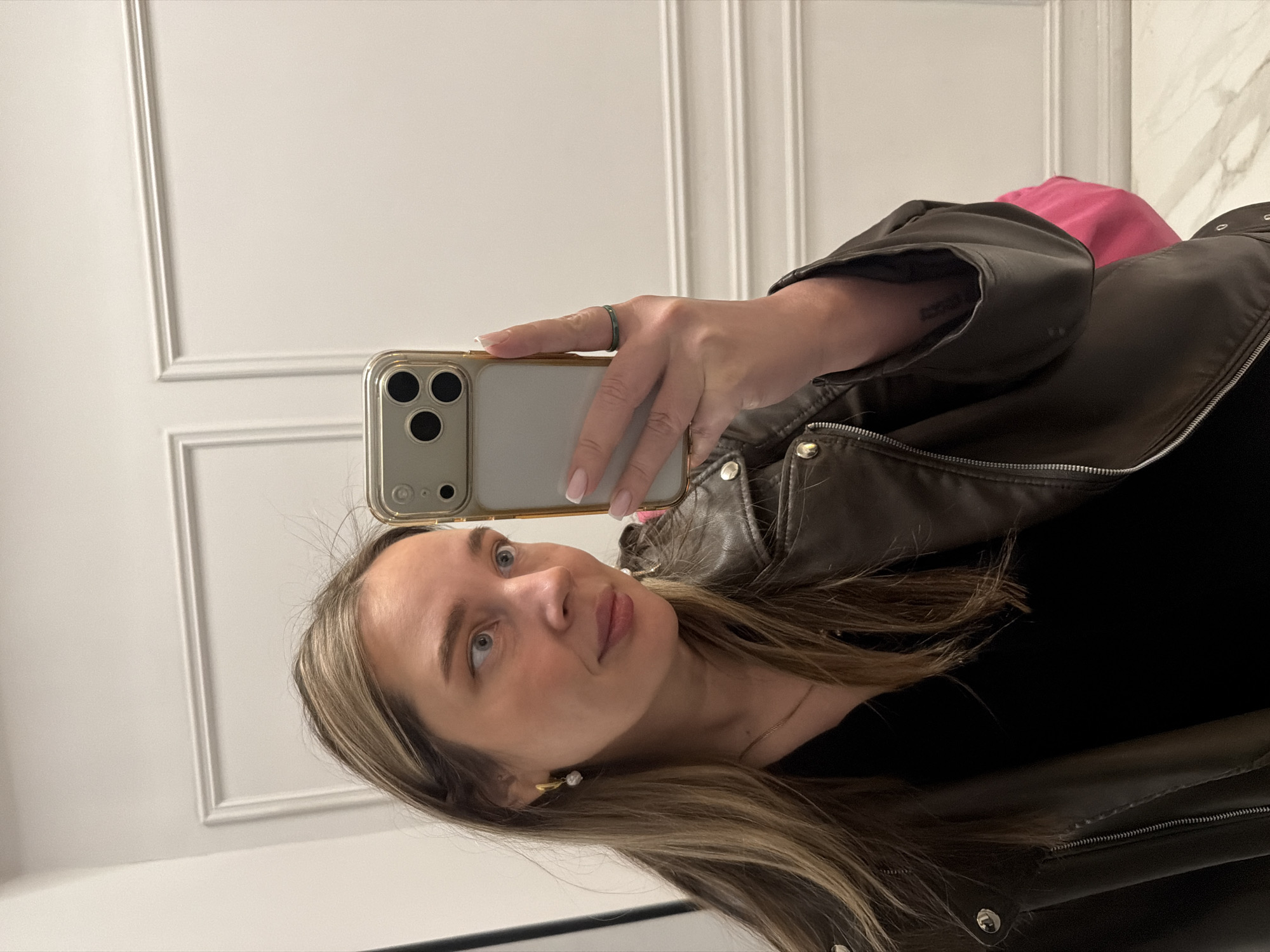 A woman is taking a selfie in a room with white walls, possibly in Moscow, Russia, though there is no definitive evidence of the location in the image. She is wearing a dark-colored shirt under a brown leather jacket. She is holding a gold-colored iPhone in her right hand. There is a pink object in the background.