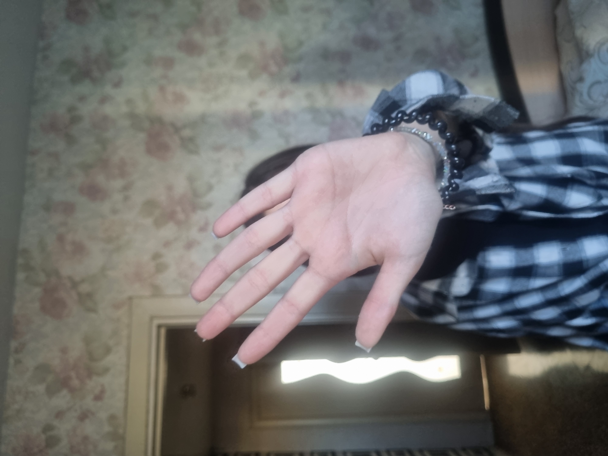 A hand extends towards the camera in front of a blurred background. The hand is fair-skinned with short, squared nails painted with a light grey or silver color. A beaded bracelet adorns the wrist, partially visible beneath the sleeve of a black and white checkered or plaid shirt.
The background features faded floral wallpaper above a doorway or a piece of furniture with a light reflecting off its surface. I cannot confirm this photo was taken in the city of Chaikovsky, Russia, due to a lack of specific identifying details in the image.